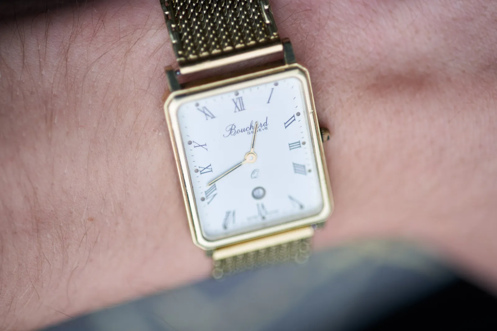 A gold watch on somebodies wrist. It seems to say Bouchard Geneve on the face but it’s not entirely clear. The hour hand covers the a of Bouchard. The clock points at 19 minutes to 1.