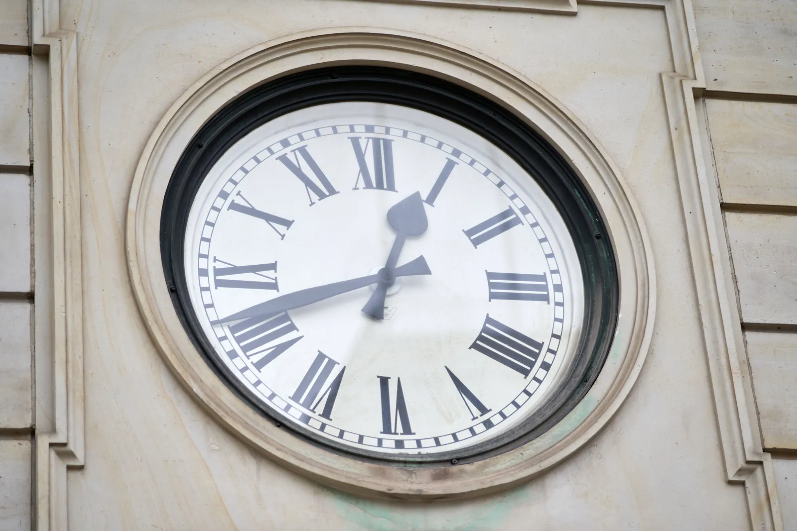 A closeup of a large clock built into a wall. The clock has roman numerals and it tells the time as 18 minutes to 1.