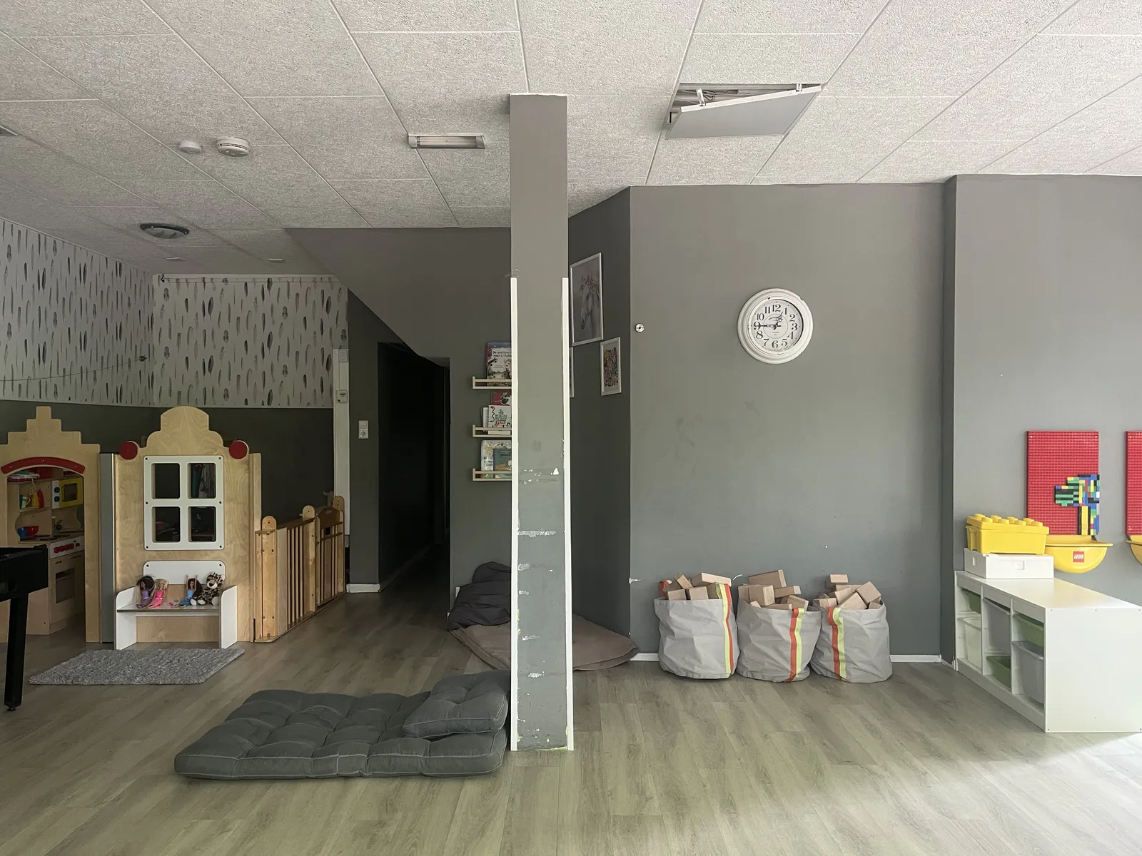 In a kindergarten with gray walls, a wooden toy house, some wooden bricks, a gray mat, and some other things for little kids. There’s a clock on one of the walls. It points at a quarter to one.