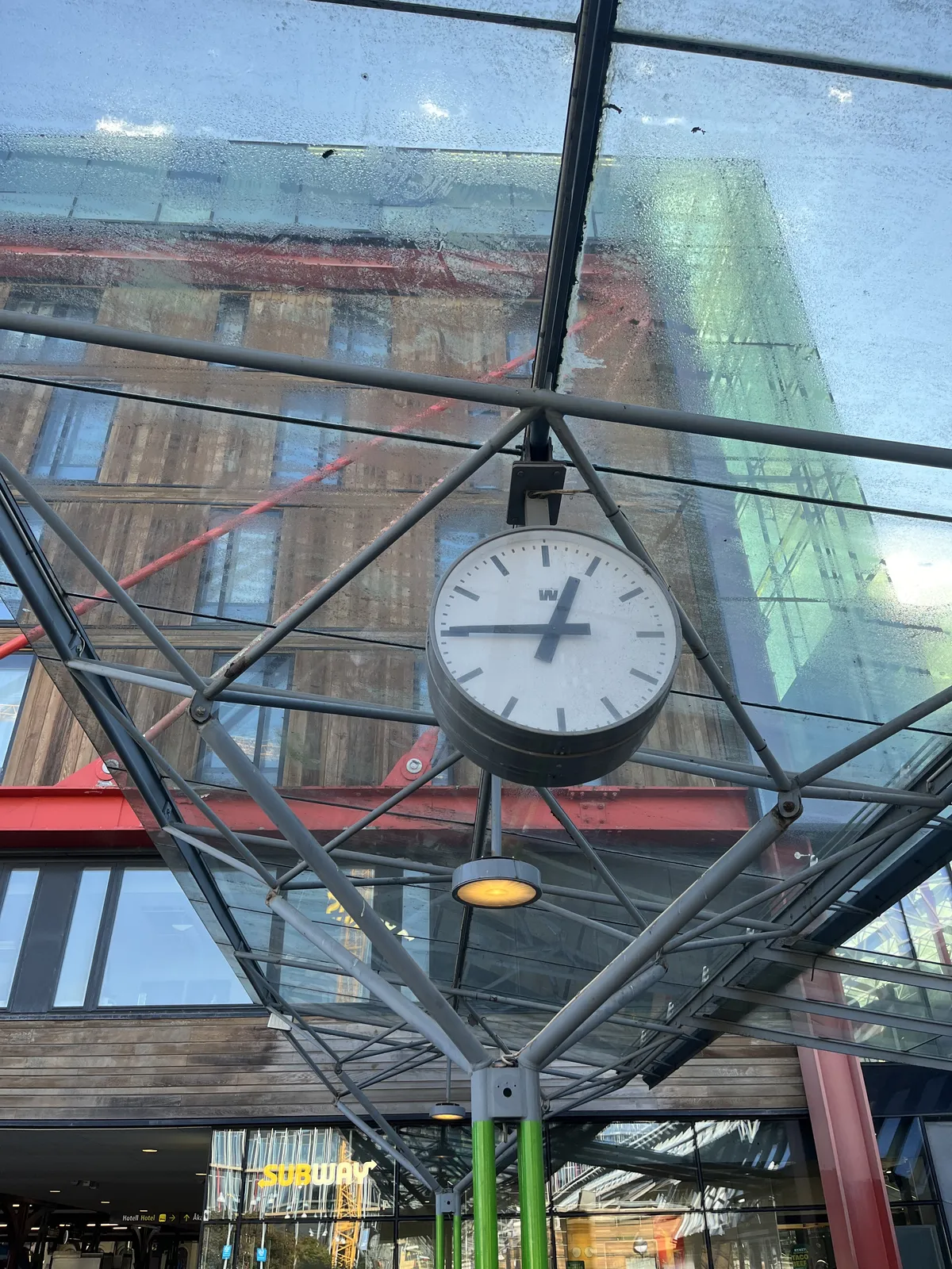 Looking up at a glass ceiling with a metal triangular construction. The glass is fogged-up, behind it, blurry, some buildings. There’s a clock hanging from the ceiling which points at a quarter to one.