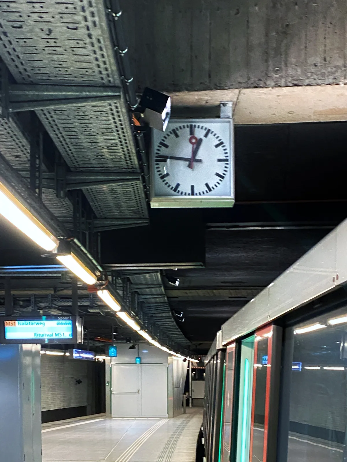 A clock attached to a concrete ceiling, next to a parked subway train. The clock points at 14 minutes to one.