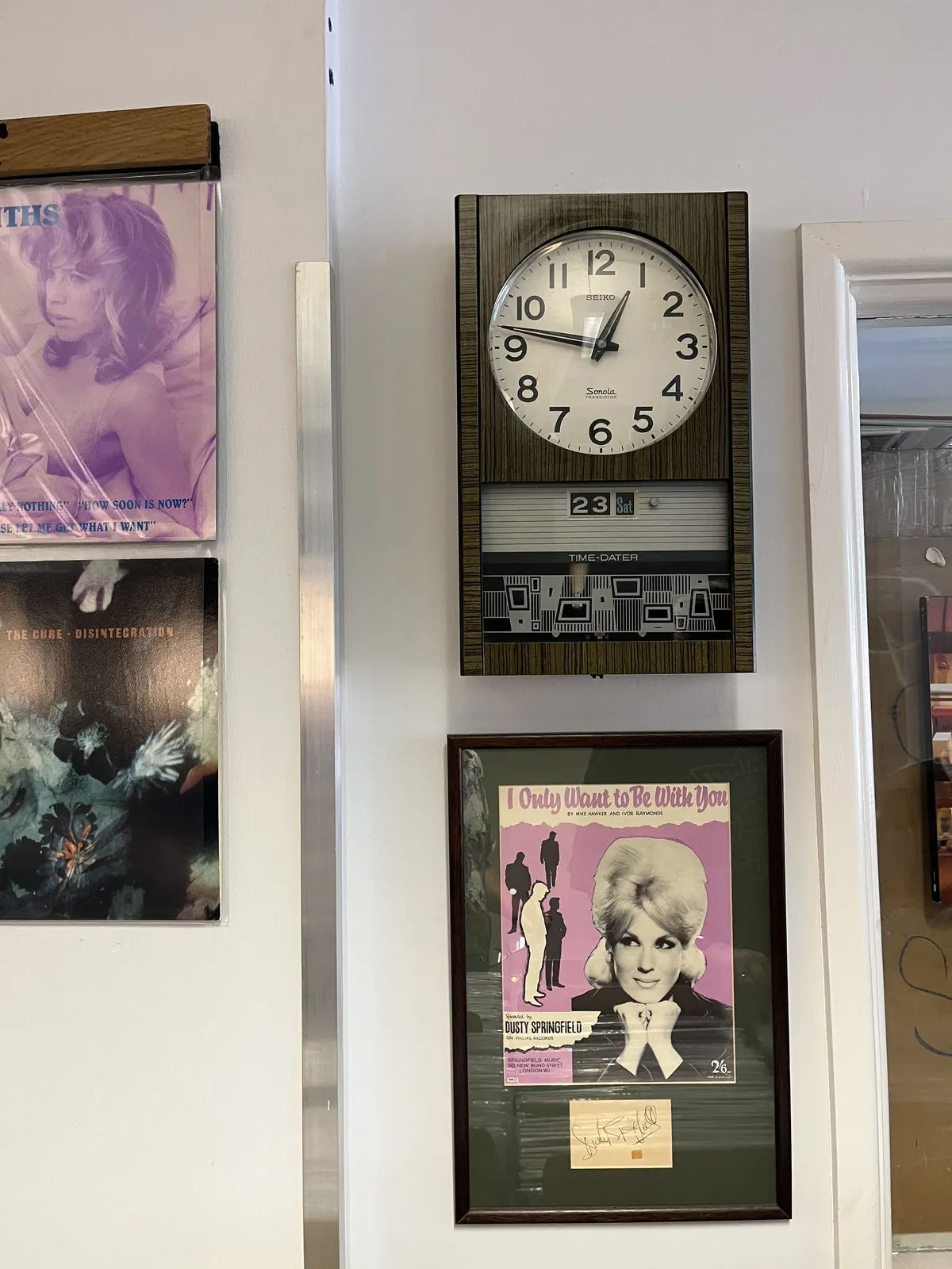 There are a few records on display on this wall, and there’s a clock. There’s a record by the Cure, and one by Dusty Springfield. On the face of the clock it says Seiko and Sonola Transistor. Below the clock it says Time Dater. The clock shows the date as 23 Sat, and the time as 13 minutes to one.