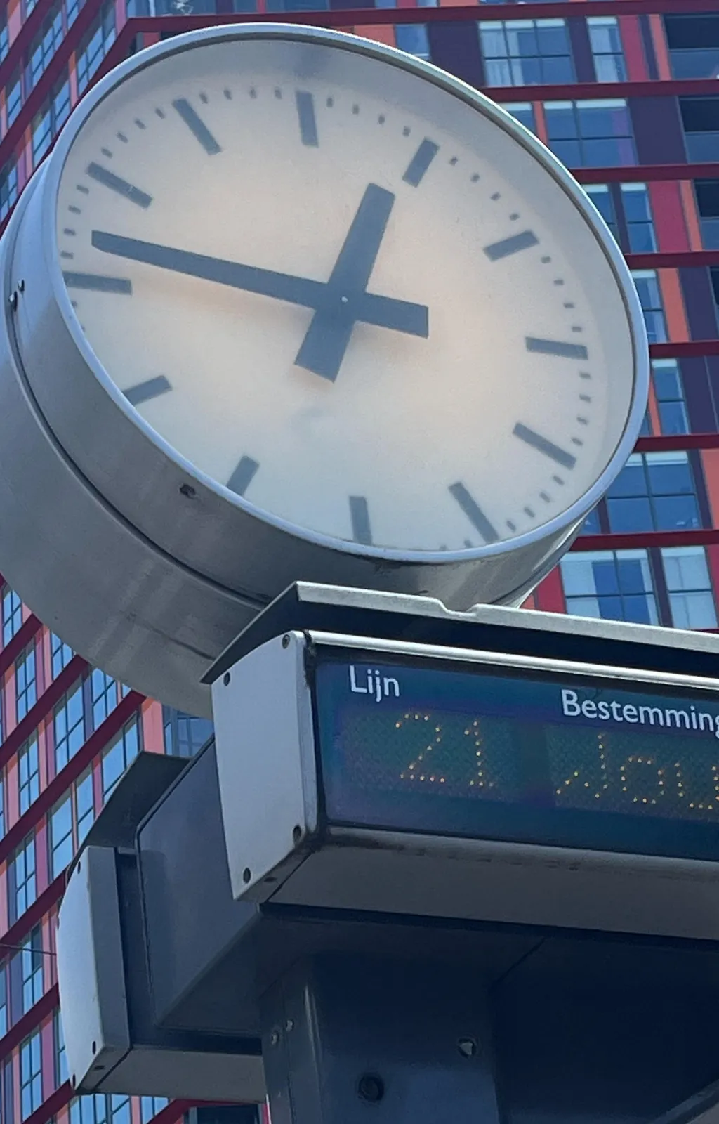 A clock on top of a sign on a tram stop in Rotterdam. In the background there’s a very large red and blue building. The clock points at 13 minutes to one.