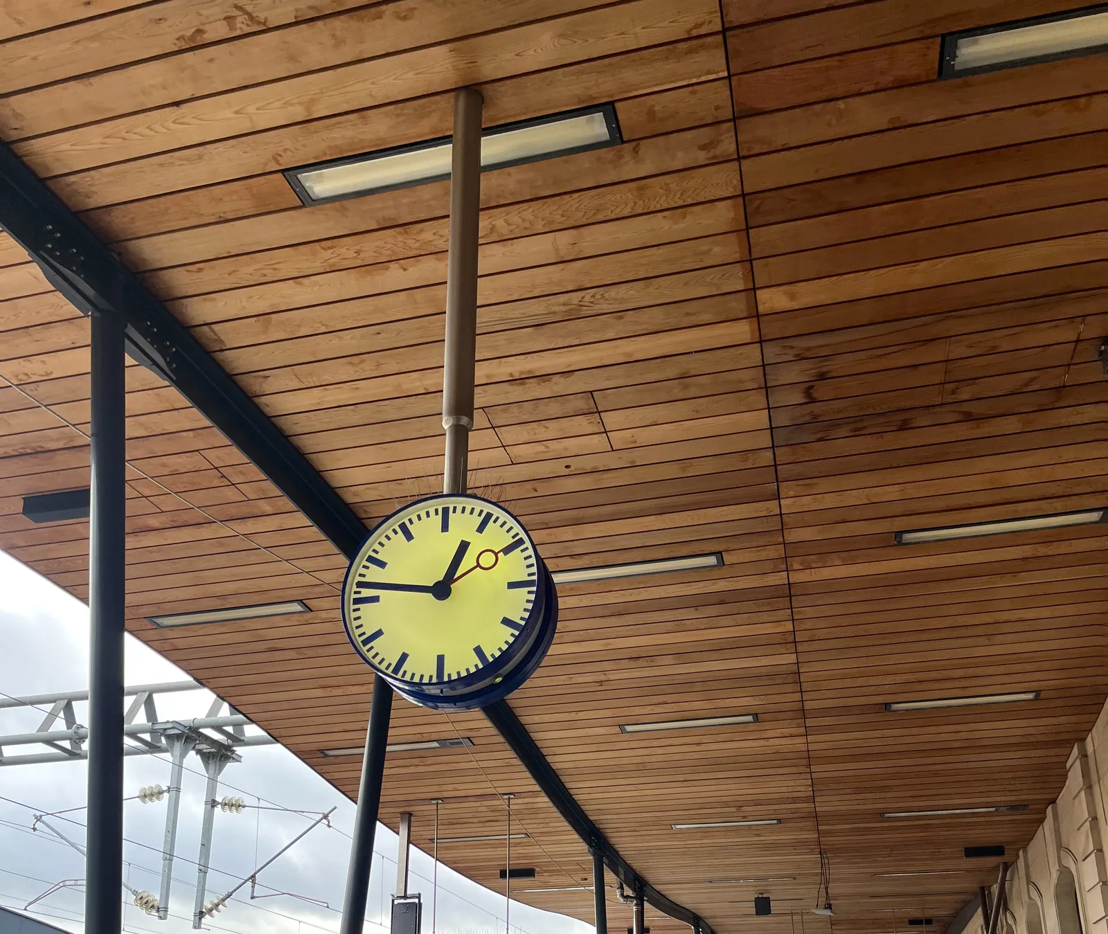 A station clock, hanging from a wooden ceiling, points at 13 minutes to one.
