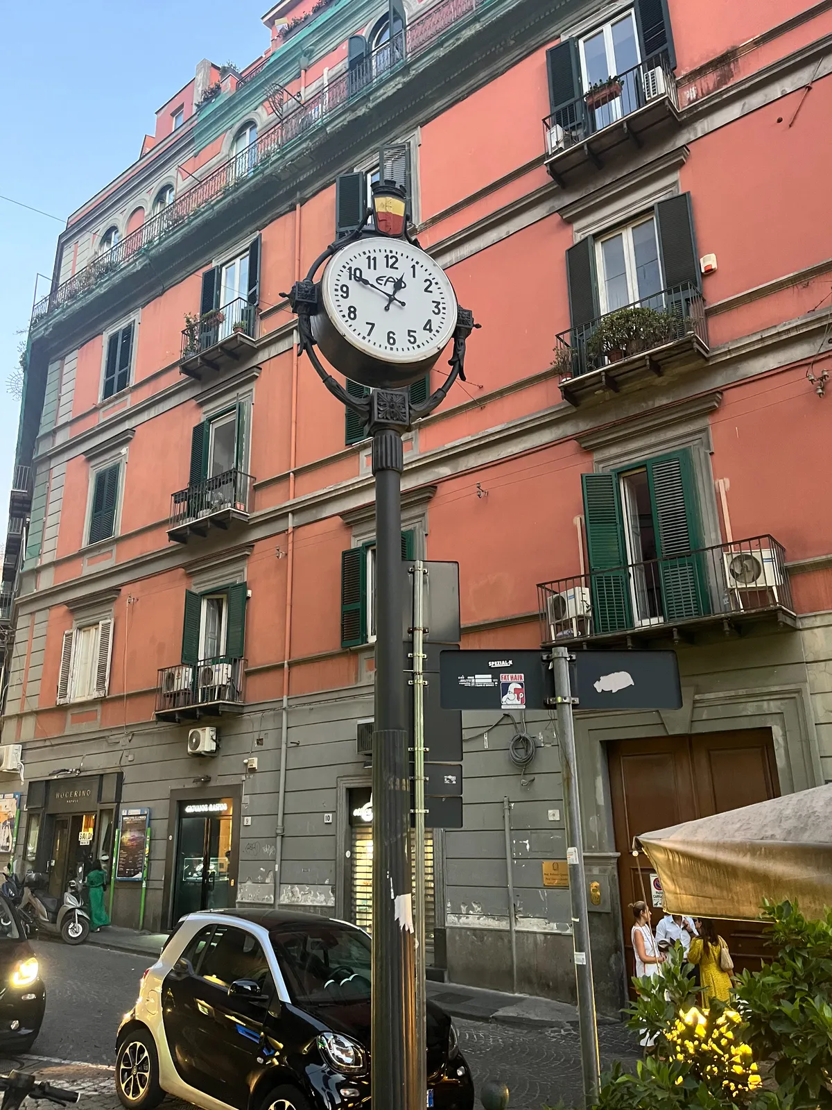 A high, peach coloured building. In front of this building there’s a clock on a high pole that points at 11 minutes to one.