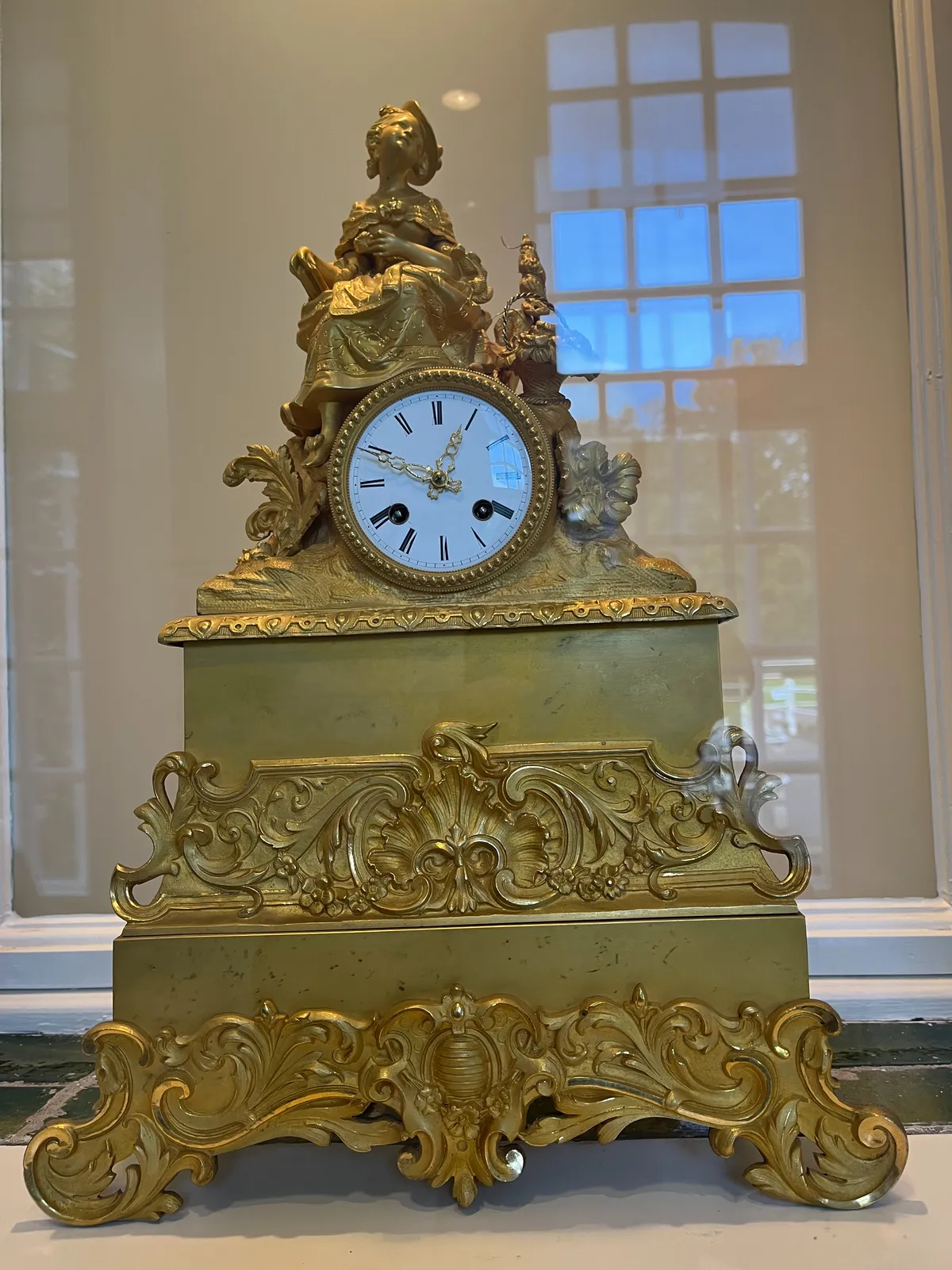 A golden, richly ornamented mantel clock with a statuette of a sitting young woman on top. It points at 11 minutes to one.