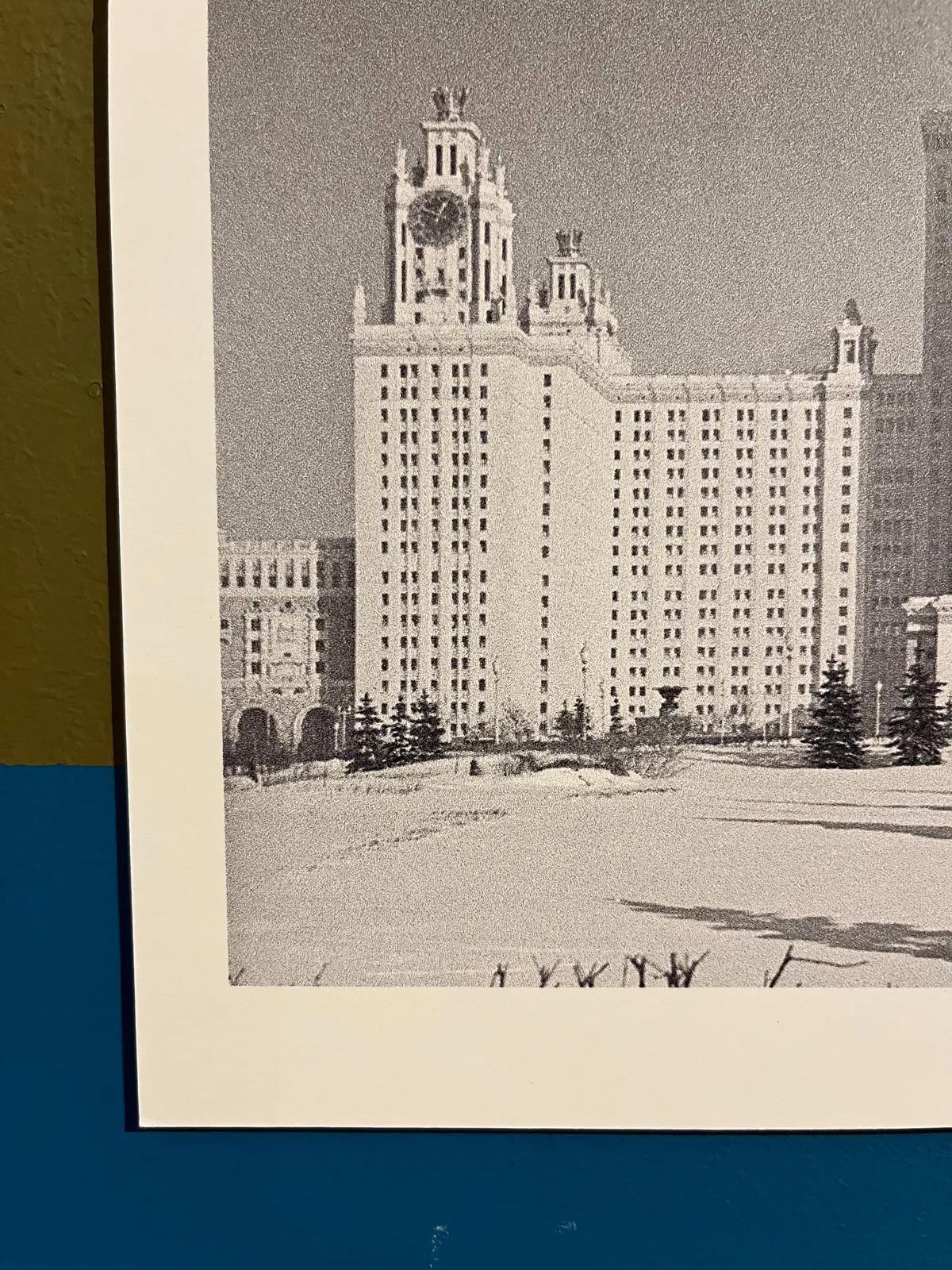 A picture of a black and white picture, pinned to a green and blue wall. On the picture is a monumental soviet building with a clock on it. It points at 10 to one.