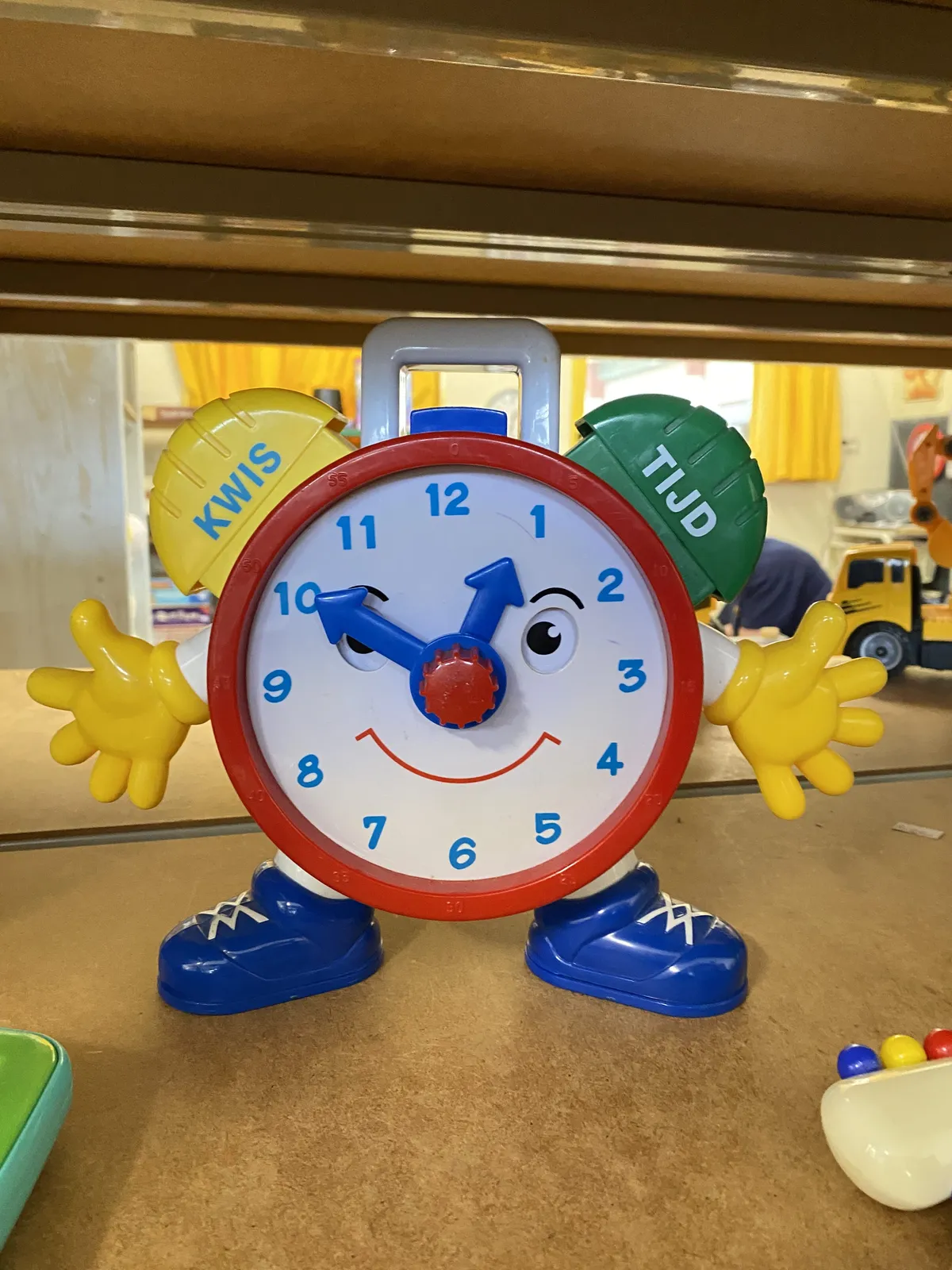 A very colourful toy clock, with a face and hands, points at ten minutes to one.