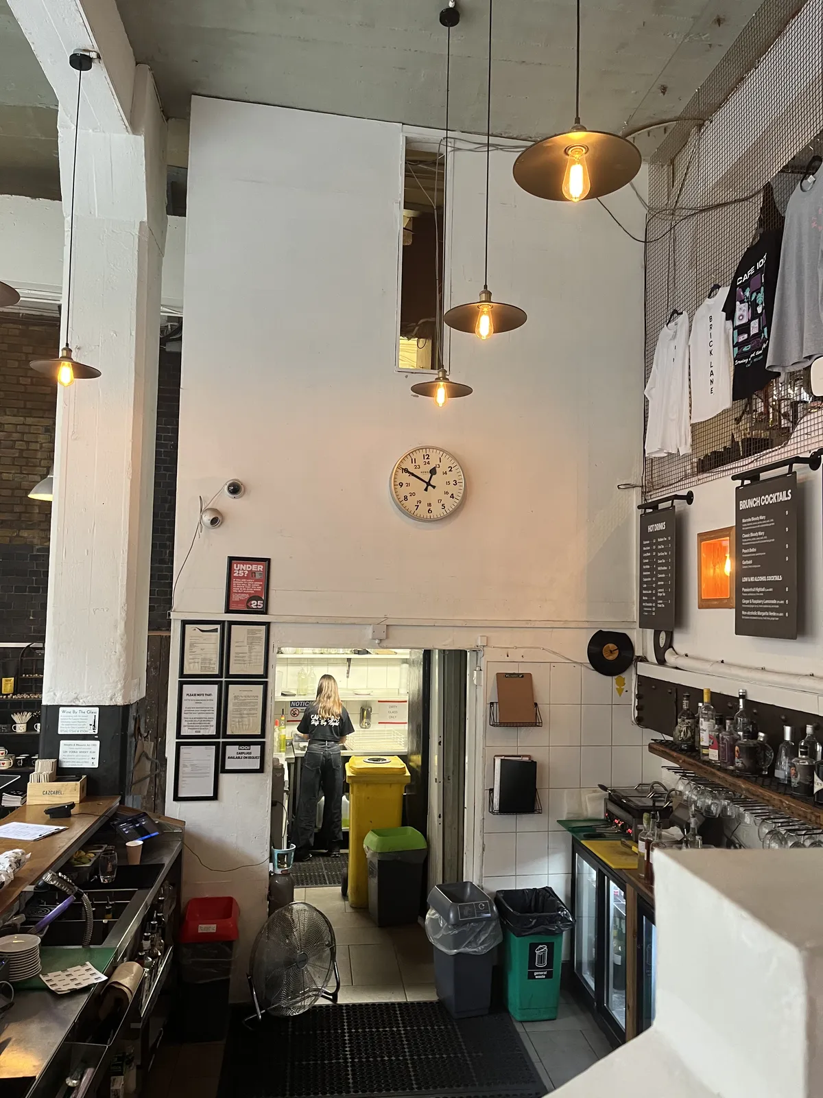 A peek behind the bar, through the door into a professional kitchen where someone is seen from behind, doing the dishes. There’s a clock on the wall above the door. It points at ten to one.