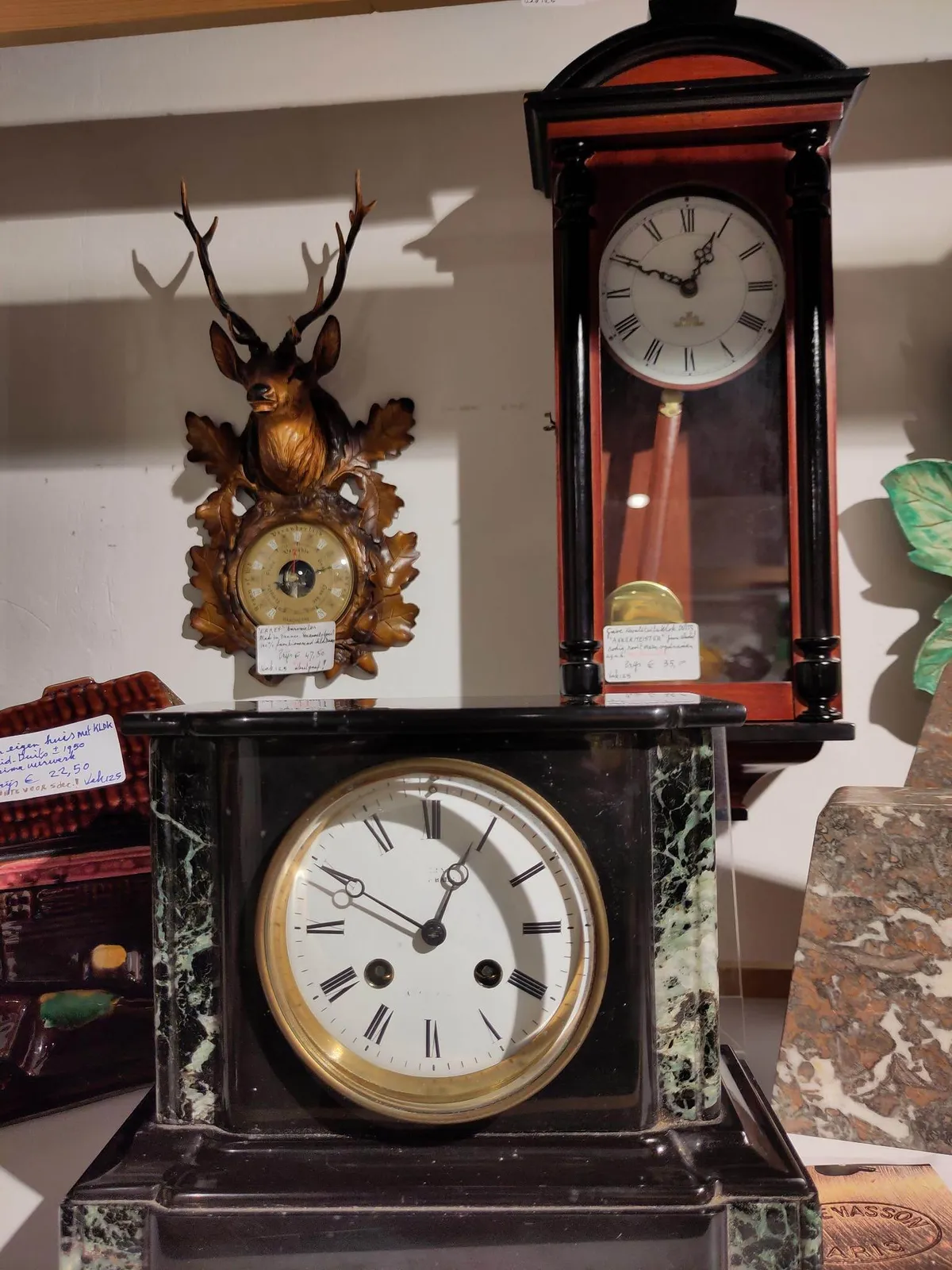 An elegant, stone mantel clock in the forground. In the background there’s an elegant, wooden wall clock. They both point at ten minutes to one.