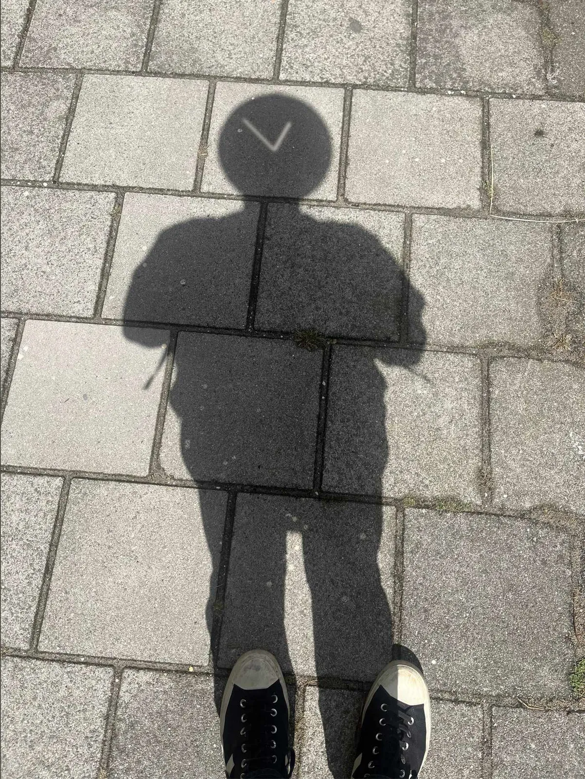 A shadow of a person on the street. The person has a remarkably round head. In this round head there are the two hands of a clock; they point at approximately 9 minutes to one.
