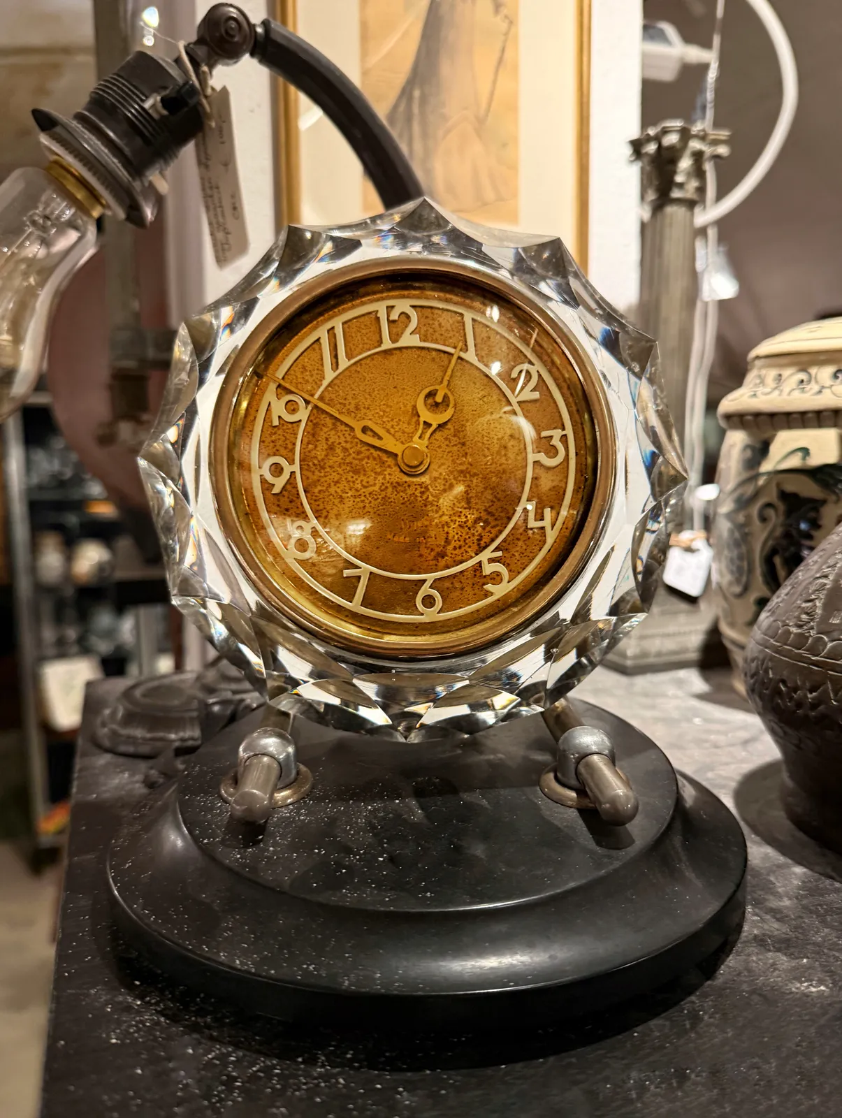 An old clock, with a rusty face, framed in a crystal frame, placed on a bakelite stand, points at 9 minutes to one.