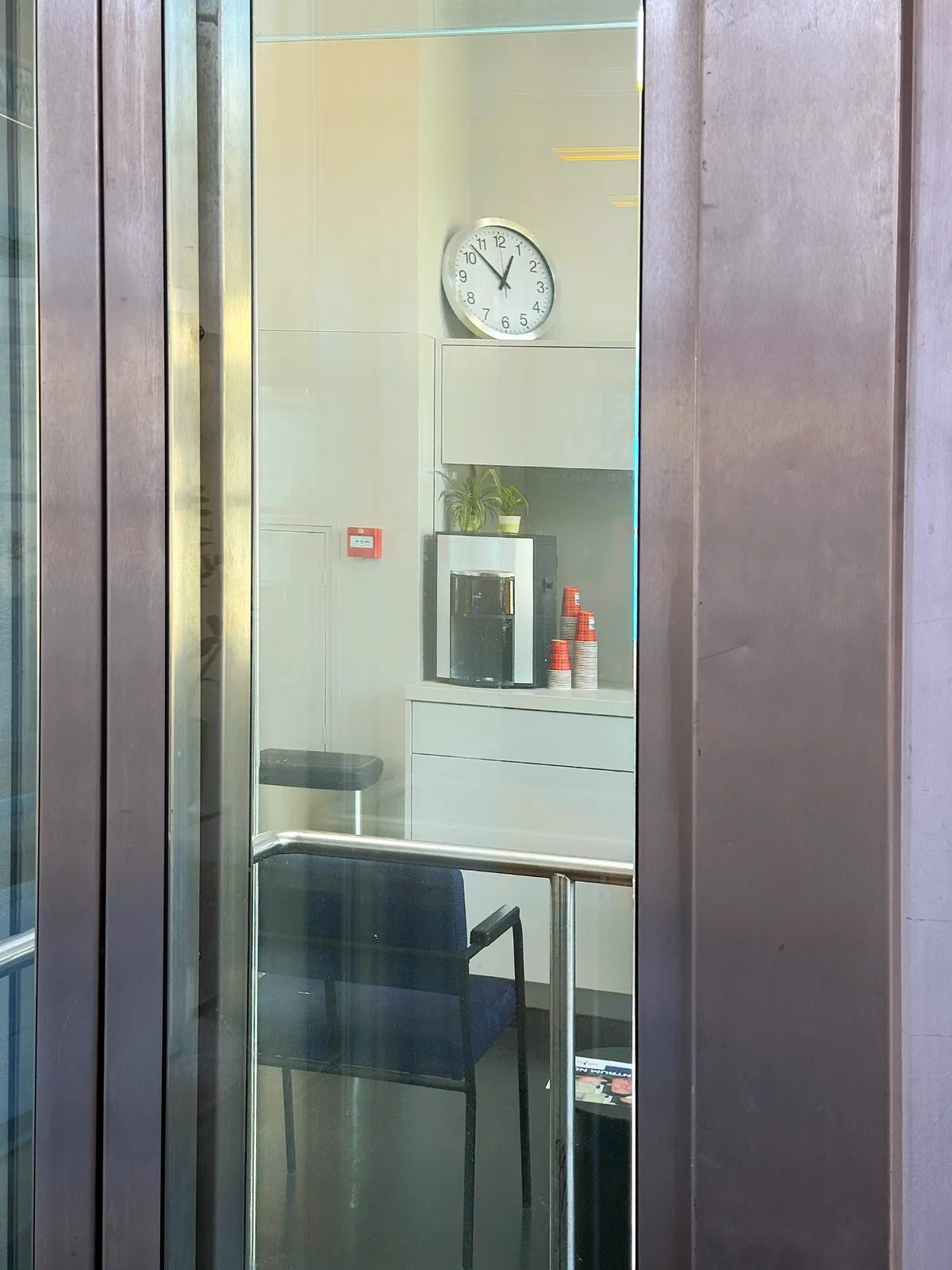 Peeking through a door into a room with a chair, a coffeemachine with paper cups, and a wall clock which points at 8 minutes to one.