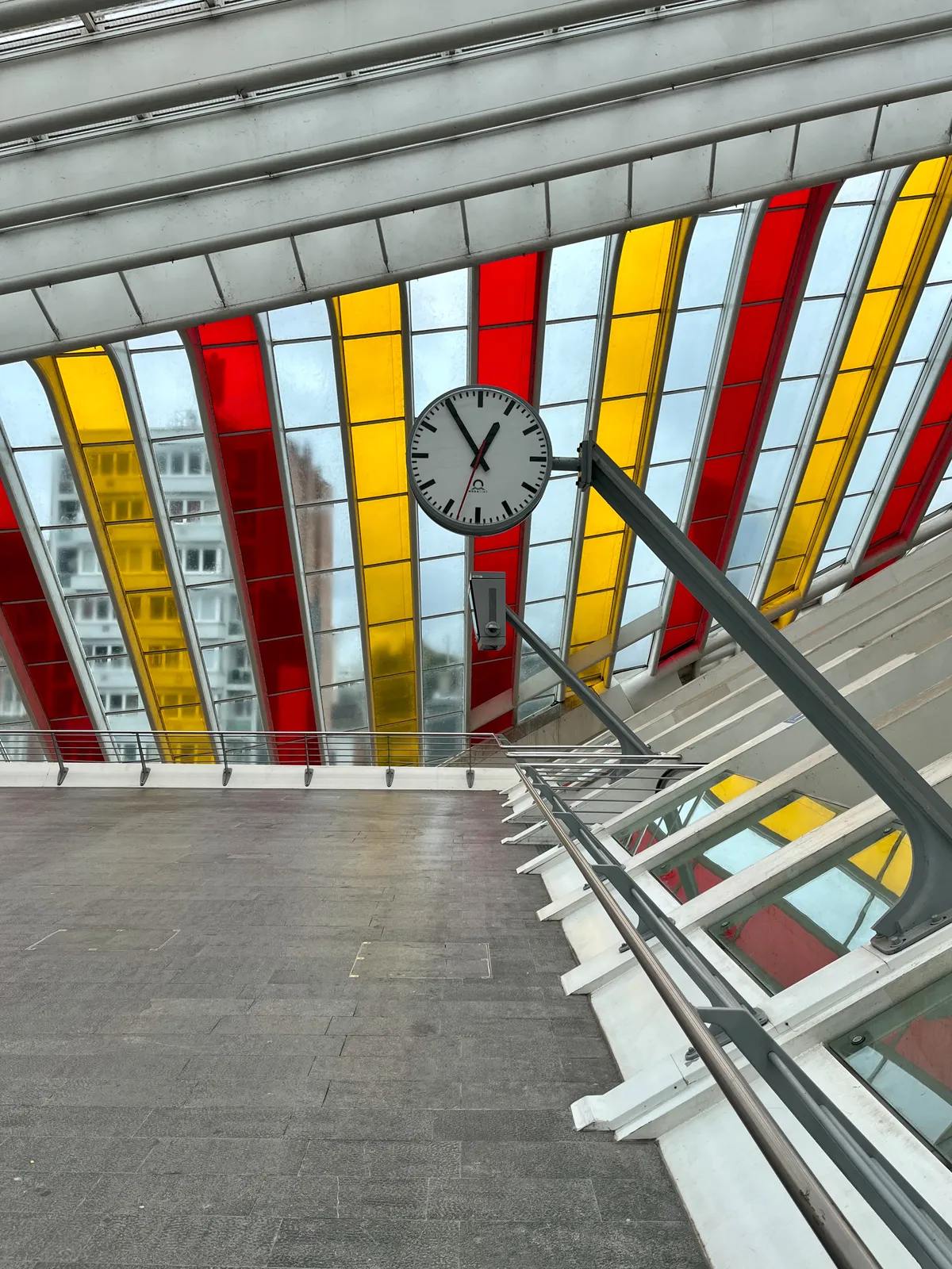 Lots of different diagonal grids that consist of white, bright red and bright orange squares. They form the dome of the Liège Guillemins train station. There’s a clock in this composition as well, attached to a diagonal pole. It points at five minutes to one. 