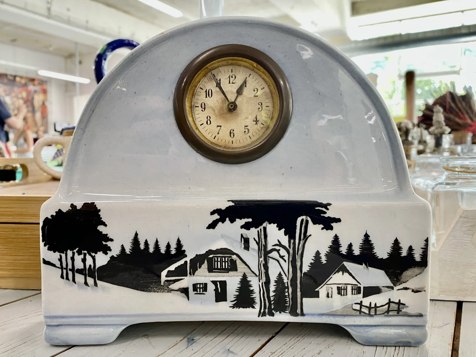 A ceramic clock. It’s a white, half circle at the top, on top of a rectangle with a house and trees painted on it, in black. On the half circle there’s an old yellowed clock that points at 5 to one.