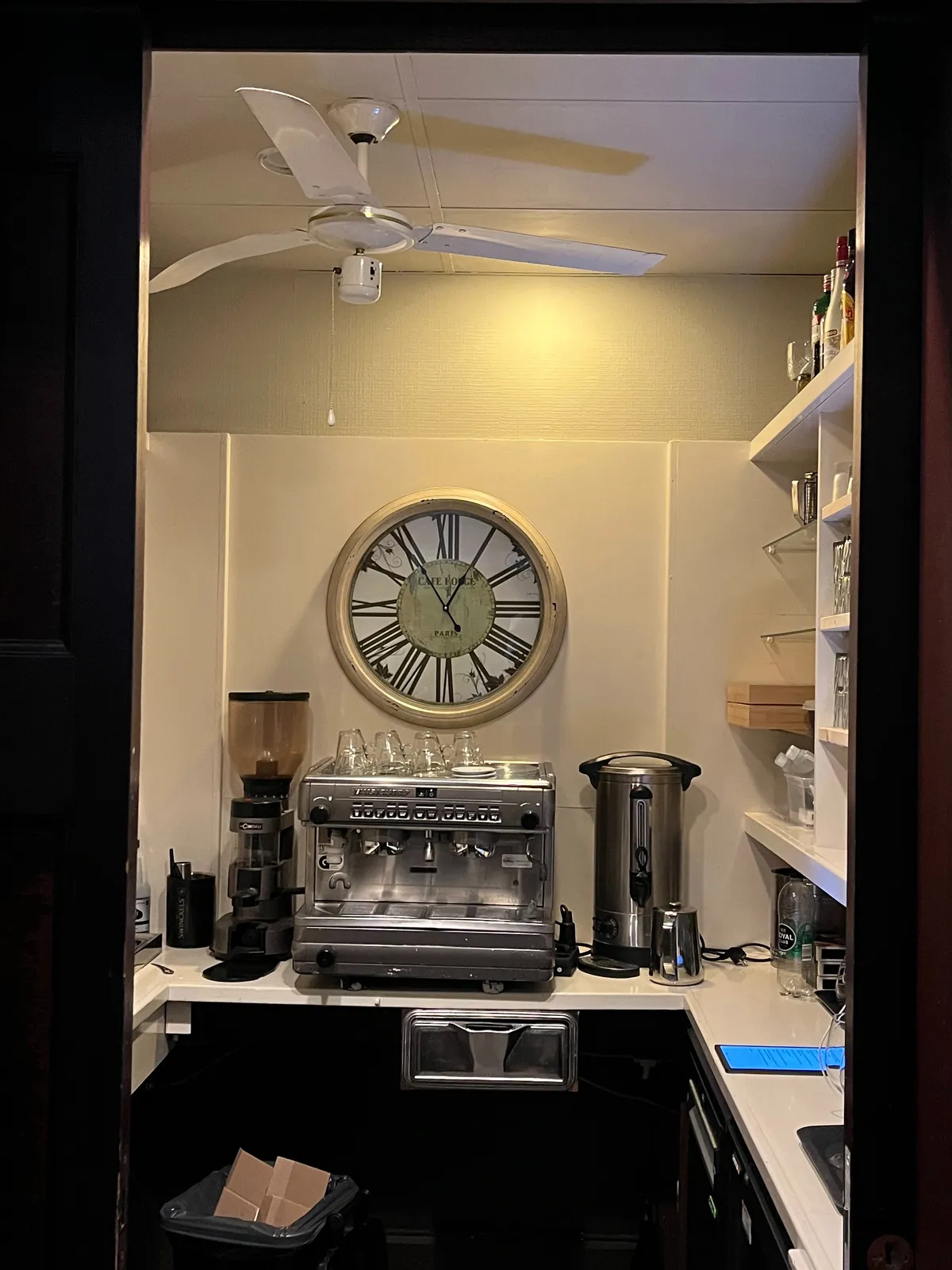Peeking through a door into a professional kitchen, with a coffee machine and a large wall clock. It points at 5 to one.