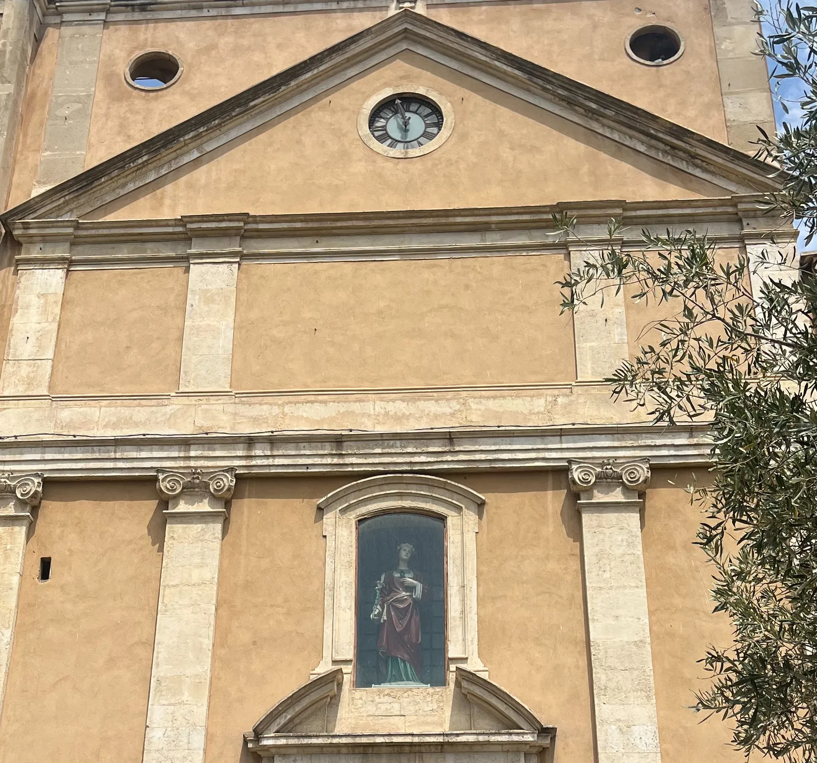 The facade of a large church. There’s a coloured statue of a holy woman behind glass. And there’s a clock. The hour hand is broken, but you can still see that it points at approximately 3 minutes to one.