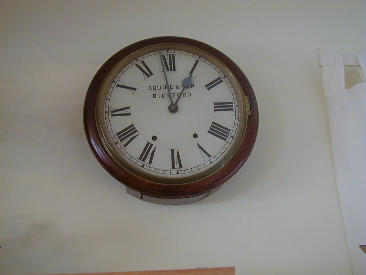 A wall clock with Roman numerals, and the text Squire & Son Bideford on its face, points at one minute to one. 