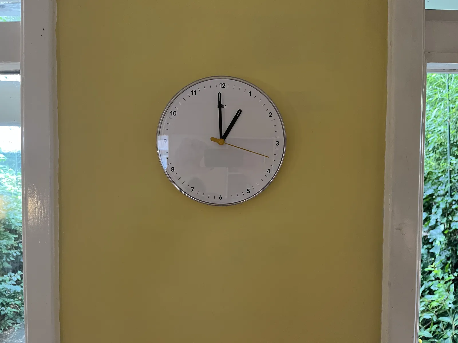On a yellow wall, surrounded by white wooden window frames, there’s a white clock, with a yellow second hand. The other hands are black. The clock points at one minute to one.
