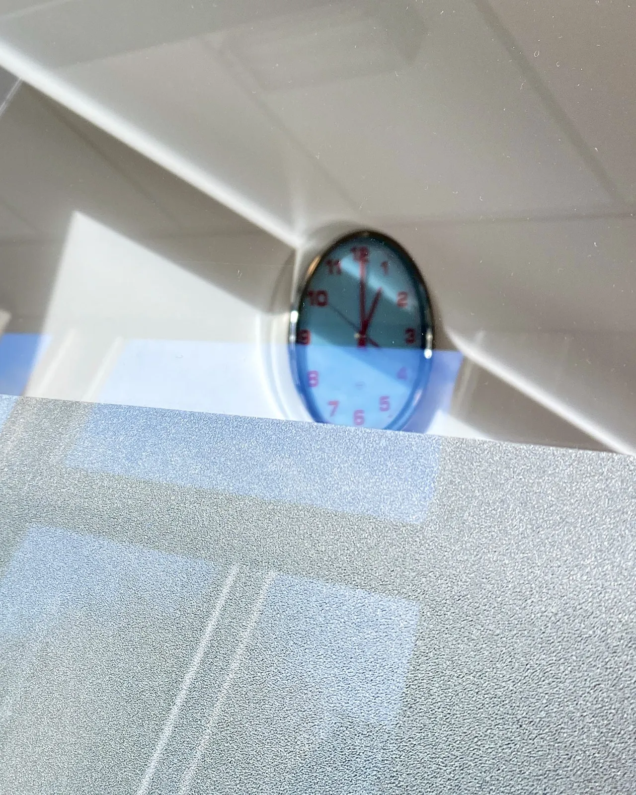 A mostly gray, almost abstract composition with a few clear diagonal lines. The lines turn out to be the ceiling and a gray wall. The wall is in focus and you can peek over it. Behind it is a clock with red hands and numbers. It is a bit blurry, but you can still see that it points at exactly one o’clock.