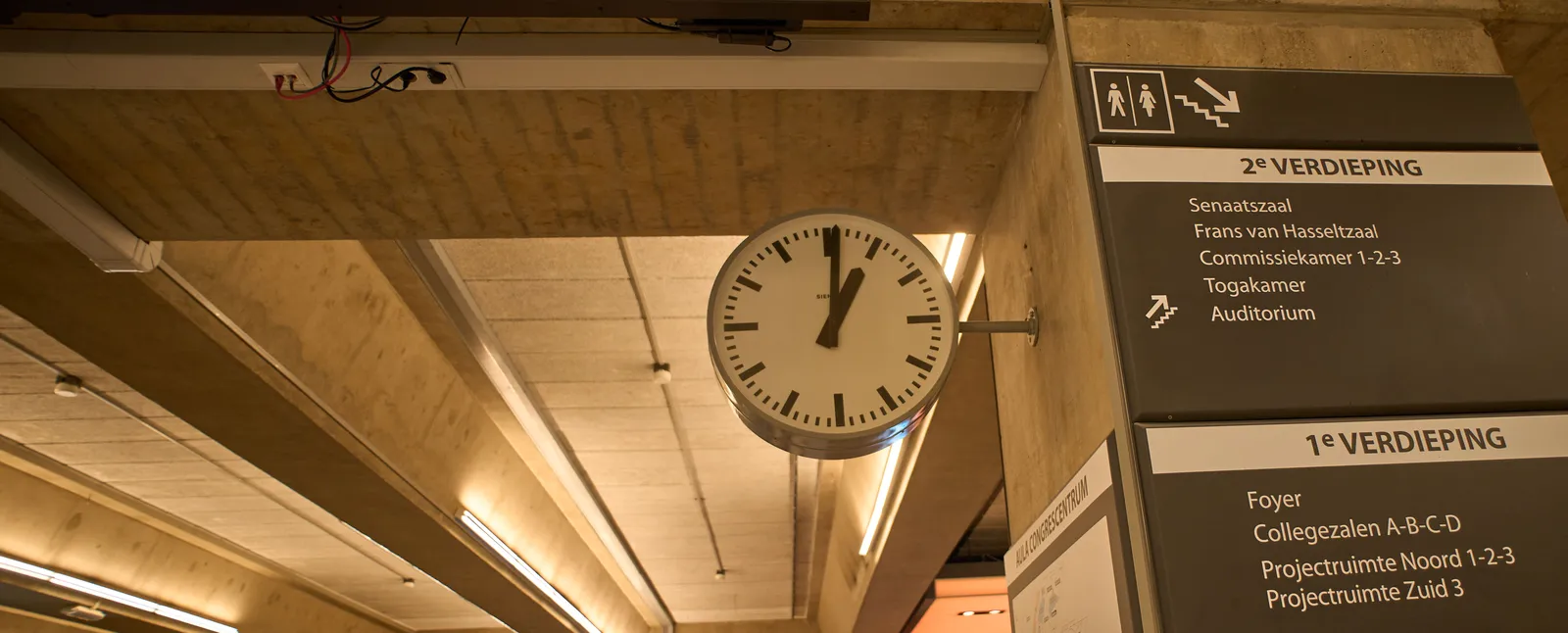 In this concrete building there are signs pointing at lecture halls and other rooms within a university. And there’s a clock which points at 1 minute past 1.