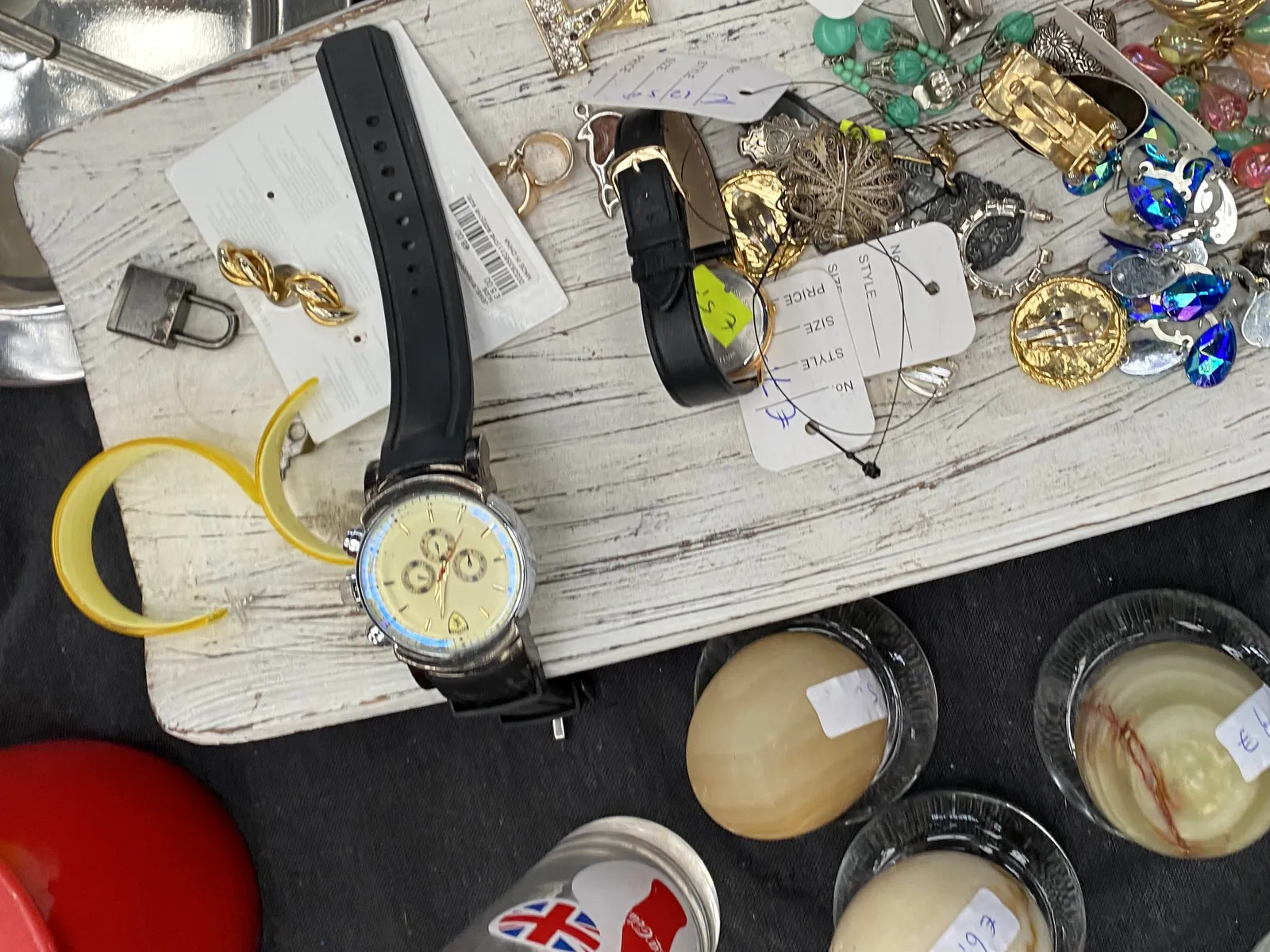 A wrist watch carelessly dropped between some other stuff in what seems to be a detail in a flea market. The watch is pictured upside down. It points at approximately 2 minutes past 1.