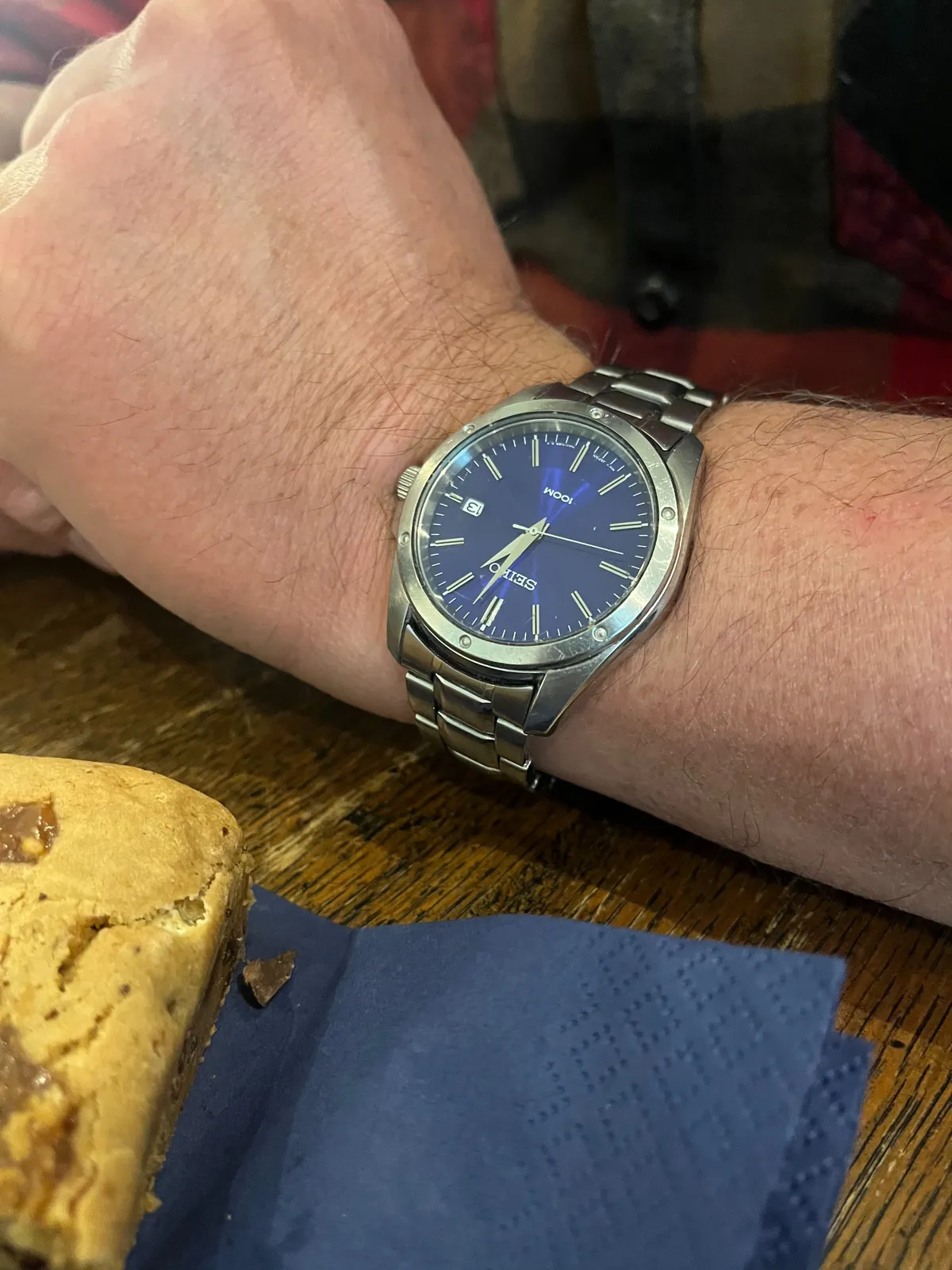 A seiko wrist watch, with a blue face, on someone’s wrist, on a table with a brownie in the foreground. The watch points at 2 minutes past one.