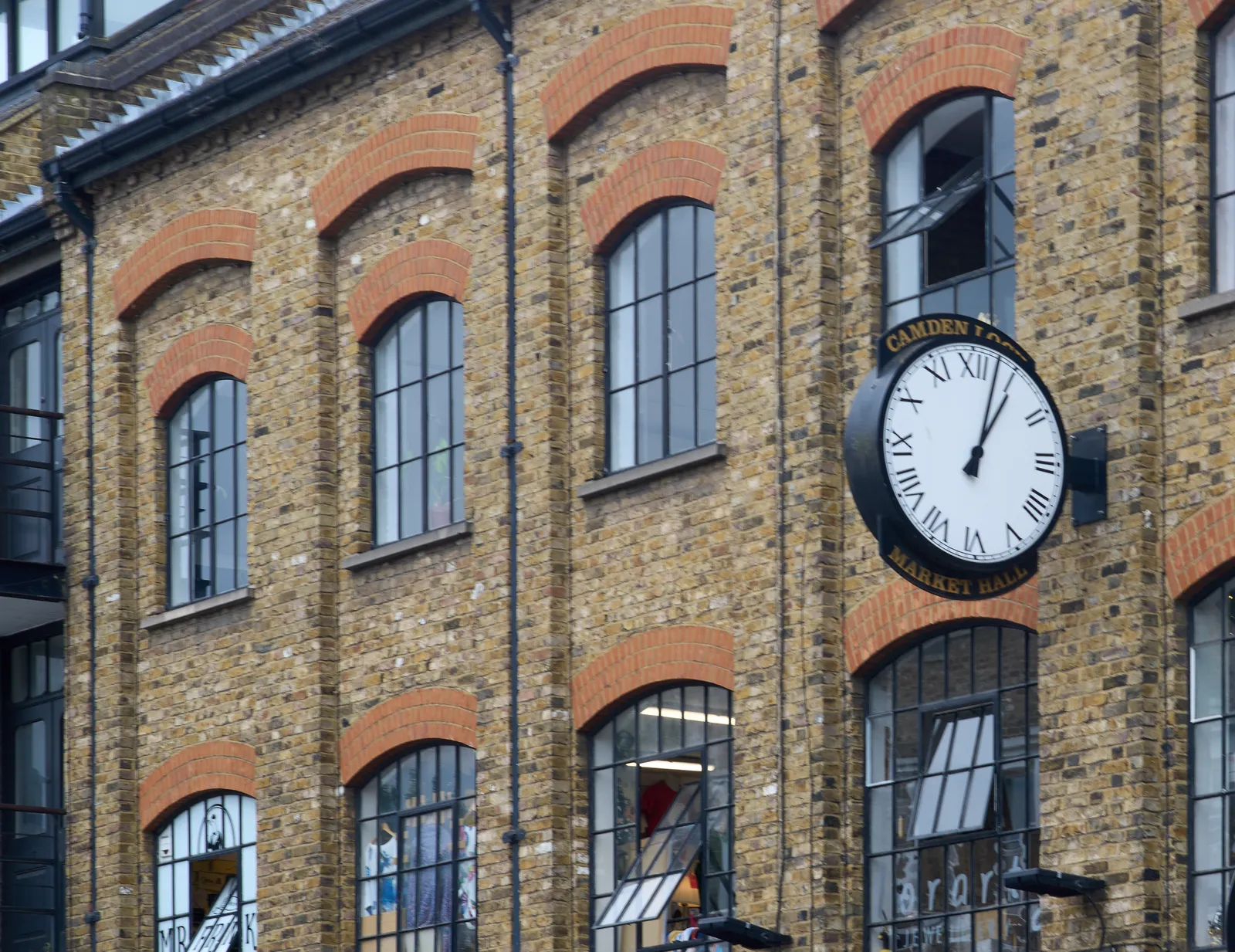 A large, black clock attached to an old, industrial, brick facade. It says Camden Lock Market Hall around the clock. It points at three minutes past one. 