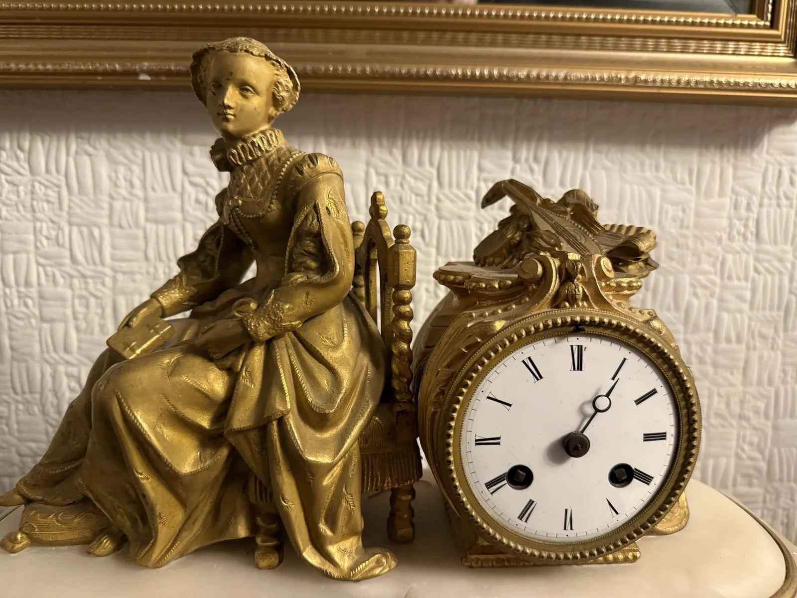 A metal statue of a lady sitting. She’s leaning against a clock. This clock misses the minute hand. But by the looks of the hour hand it is approximately 5 past one.
