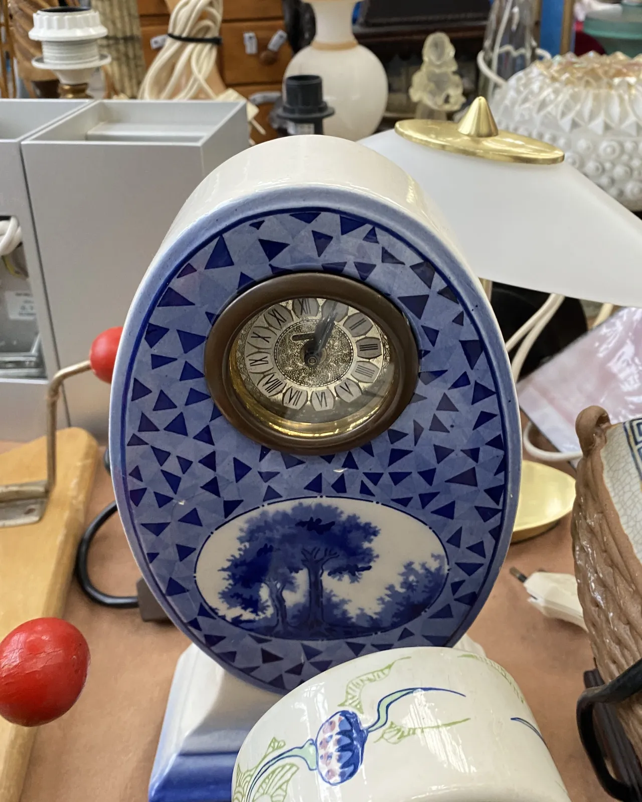 An oval, blue and white ceramic clock on a flea market points at five minutes past 1.