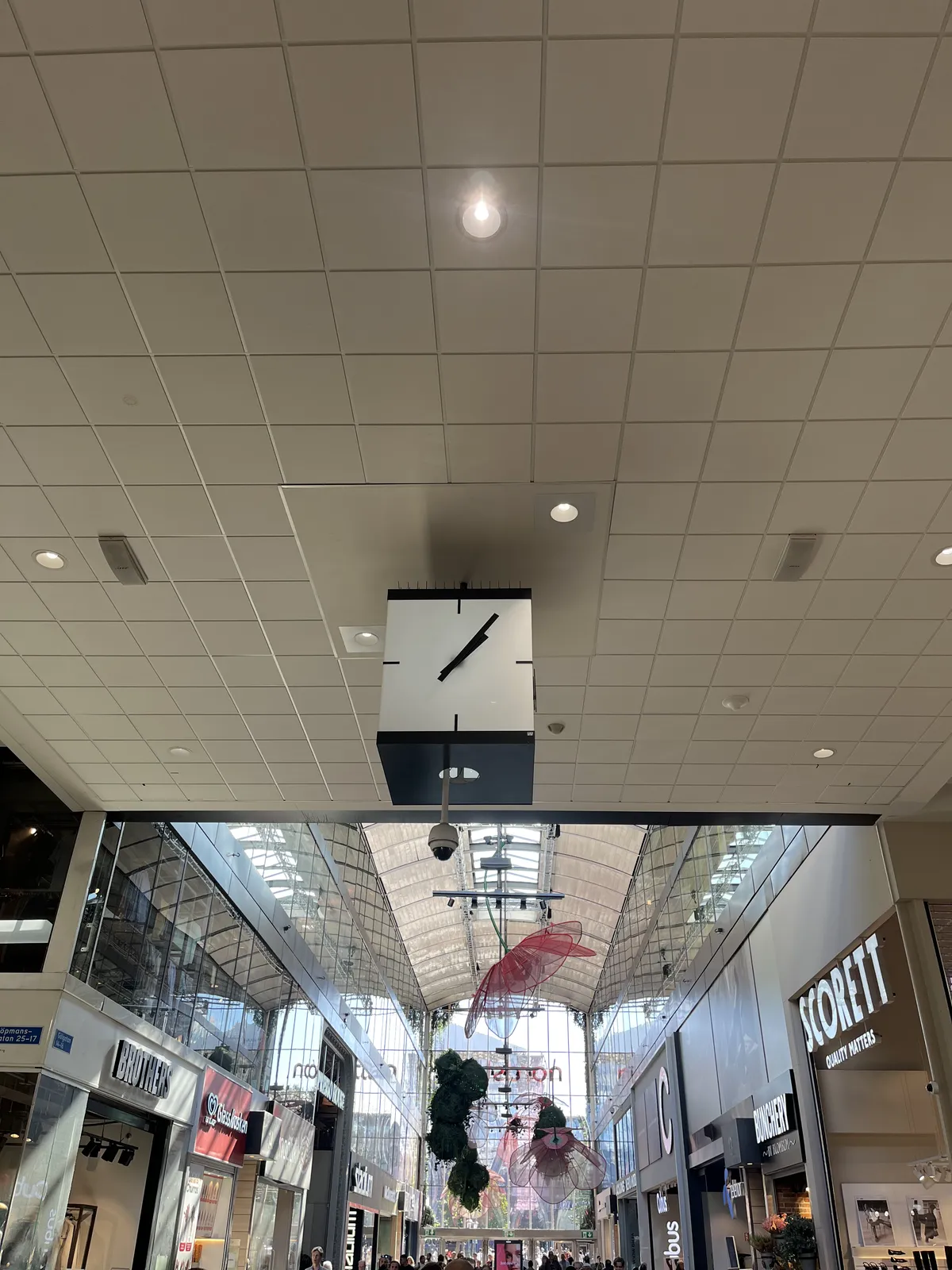 A square, minimalist clock is attached to the high ceiling of a standard shopping mall. It points at 5 past one.