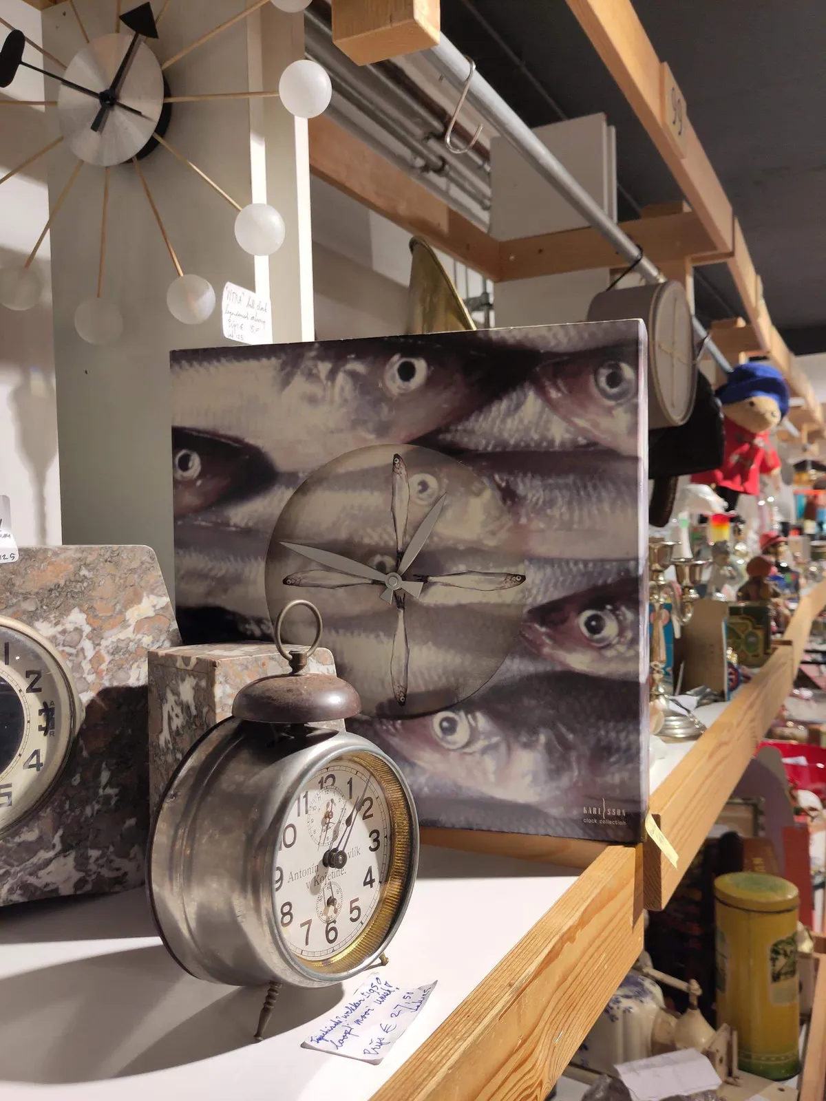 There are a few clocks in this picture. A clock made from a canvas with prints of fish on it. The hands and the indicators are fish-shaped as well. It points at 14 minutes to one. There is a clock on the wall made from spokes with white balls on the end. It points at approximately 11 minutes to one. And then there’s an old alarm clock, with one single bell on top, that points at approximately five past one.