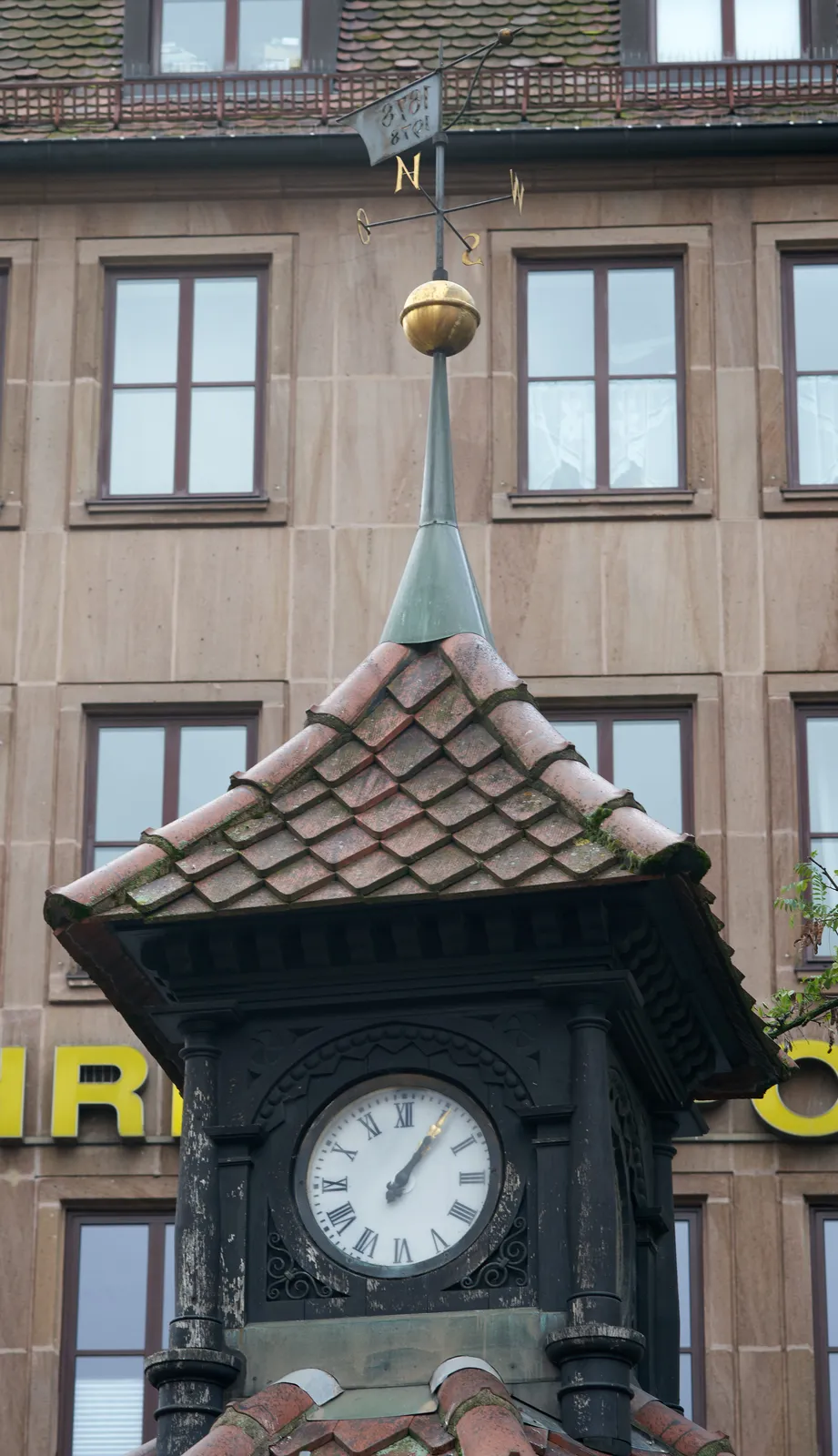 A little tower, with a much larger building in the background. On the little tower there’s a clock. It points at 5 past 1.