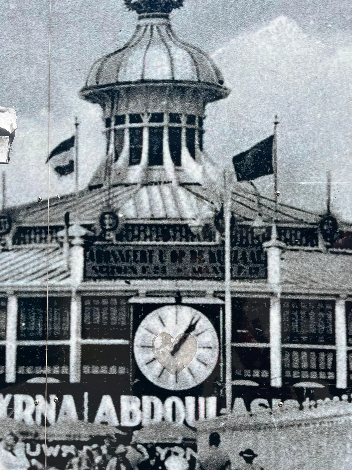 This is a picture of a picture. A heart shaped sticker has been removed from this print, you can still see the residue. On this picture there’s a clock. It points at approximately 6 minutes past 1.