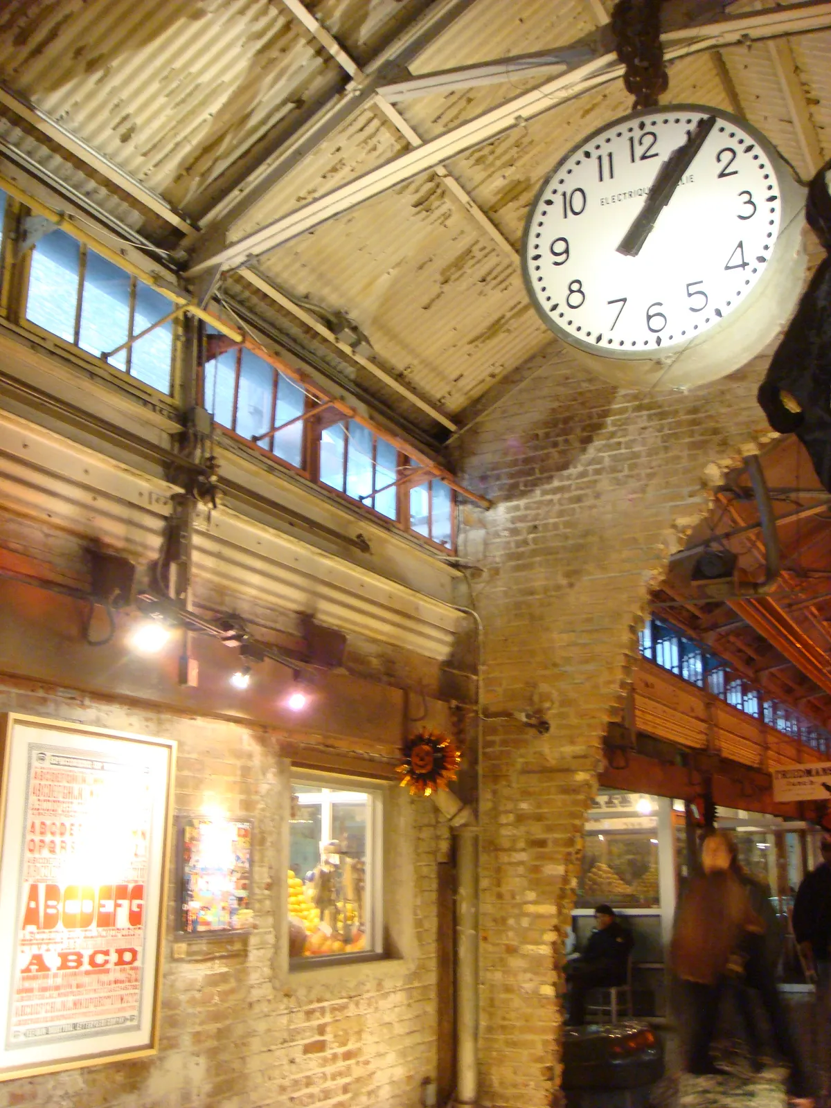 Inside an old industrial building. Brick walls, metal ceiling. There’s an old clock hanging from the ceiling. It points at 6 minutes past one.
