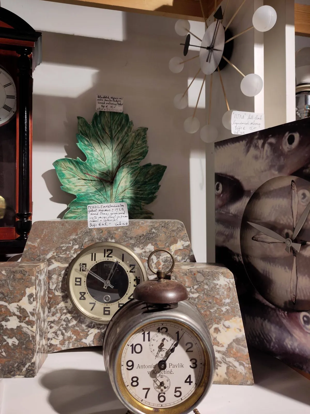 There are a few clocks in this picture. A clock made from a canvas with prints of fish on it. The hands and the indicators are fish-shaped as well. It points at 13 minutes to one. There is a clock on the wall made from spokes with white balls on the end. It also points at approximately 13 minutes to one. There’s a wall clock, in the shape of an oak leaf, that also points at 13 to one. There’s an old alarm clock, with one single bell on top, that points at approximately six minutes past one. And then there’s a classic stone mantel clock that points at ten to one.