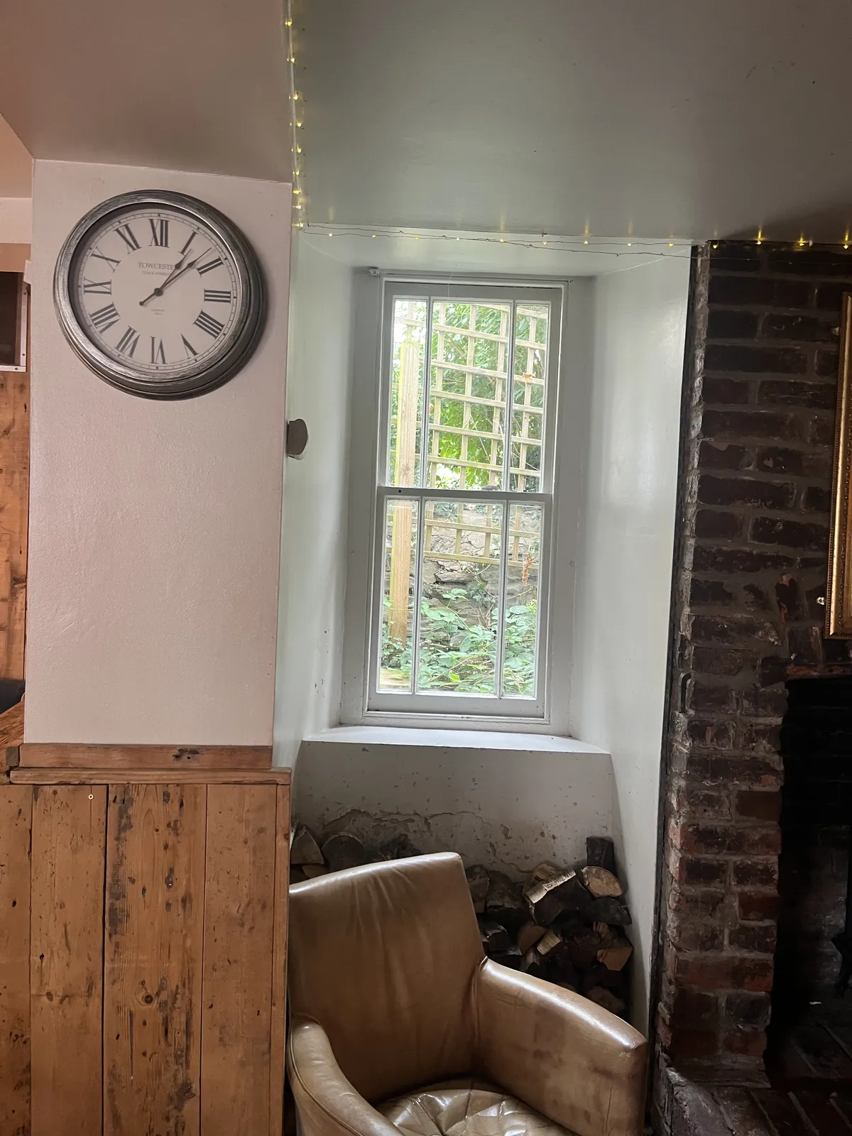 A clock on a wall. Below the clock there’s a comfy chair. Behind the chair there are some logs for a fire, and a window. The clock points at seven minutes past one.