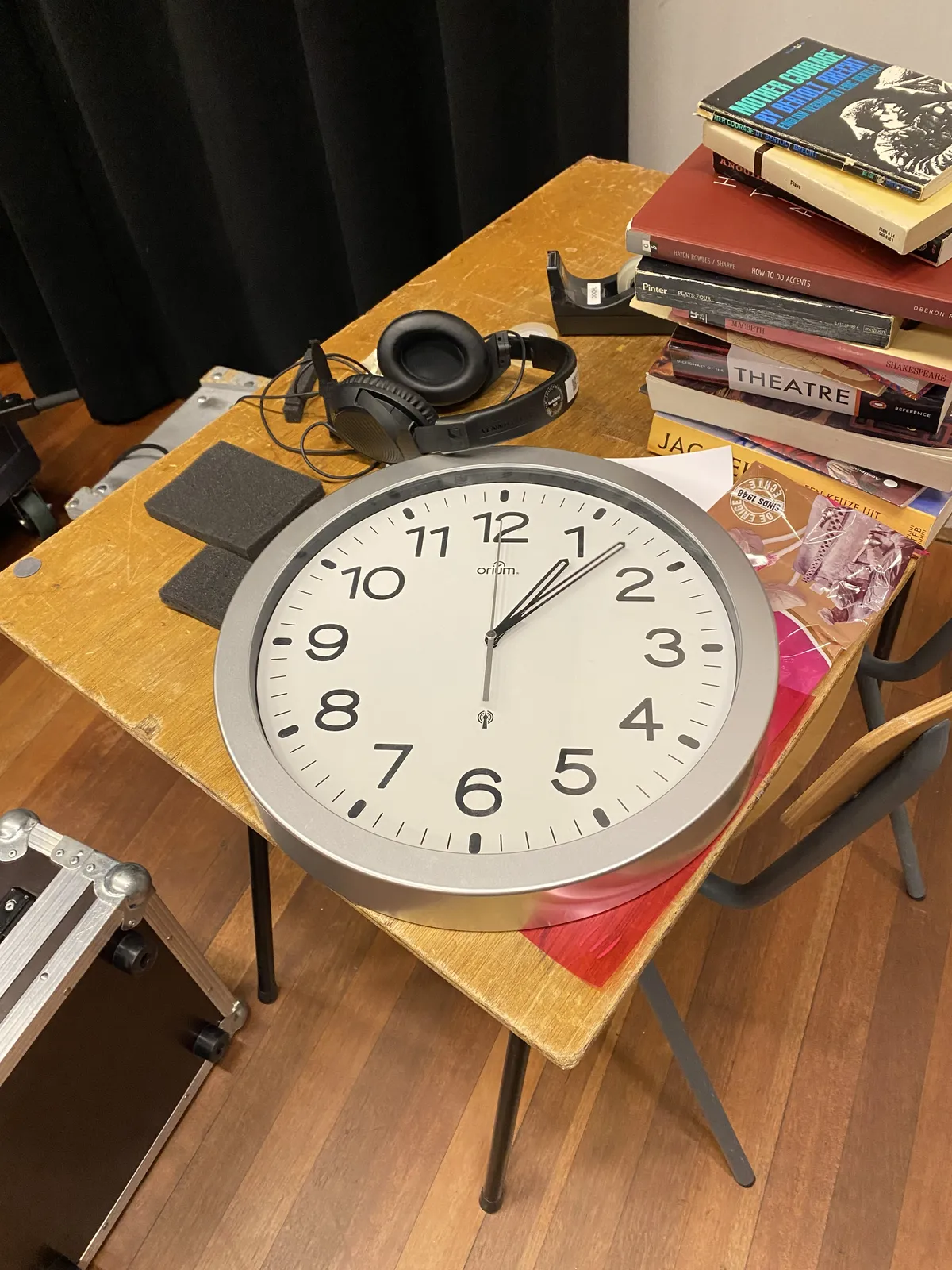 A pile of books, a headphone and a clock on a table. The clock points at 7 minutes past one.