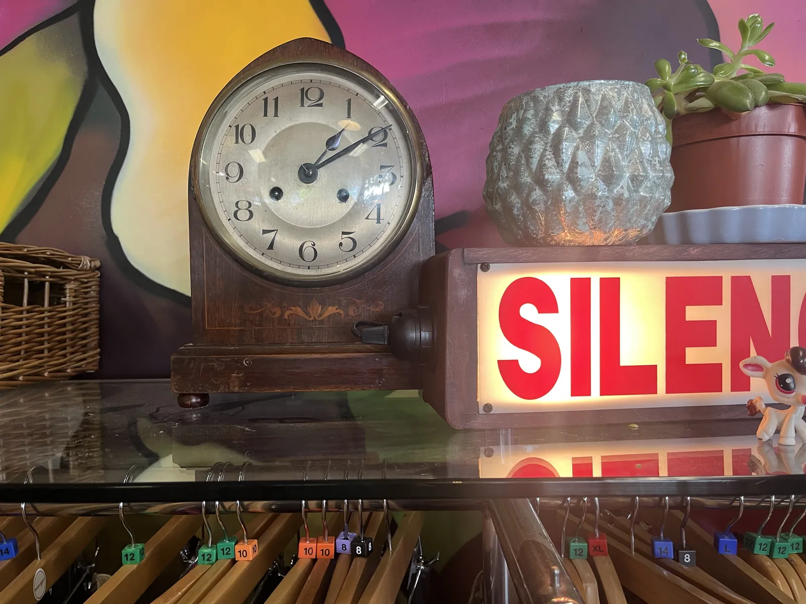 A wooden mantel clock on a glass shelf in a thrift store. There’s a lit sign next to it which probably says Silence in red letters, but only the first five letters are in the frame. There’s a colourful illustration behind the clock. It points at 9 minutes past one.