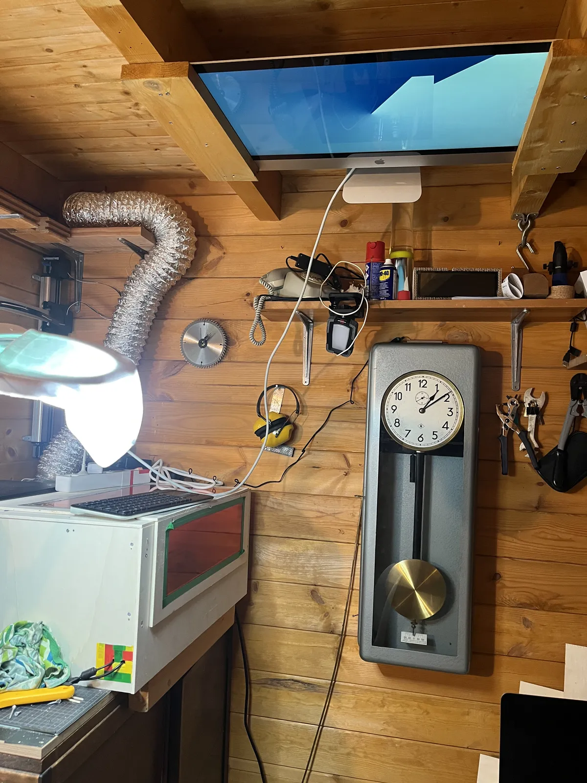 On the wall in a wooden shed there’s a mechanical clock, surrounded by tools. There’s a large computer screen attached to the ceiling with an abstract shape on it, which resembles a clock. Both the abstract clock and the mechanical clock point at 9 minutes past one.