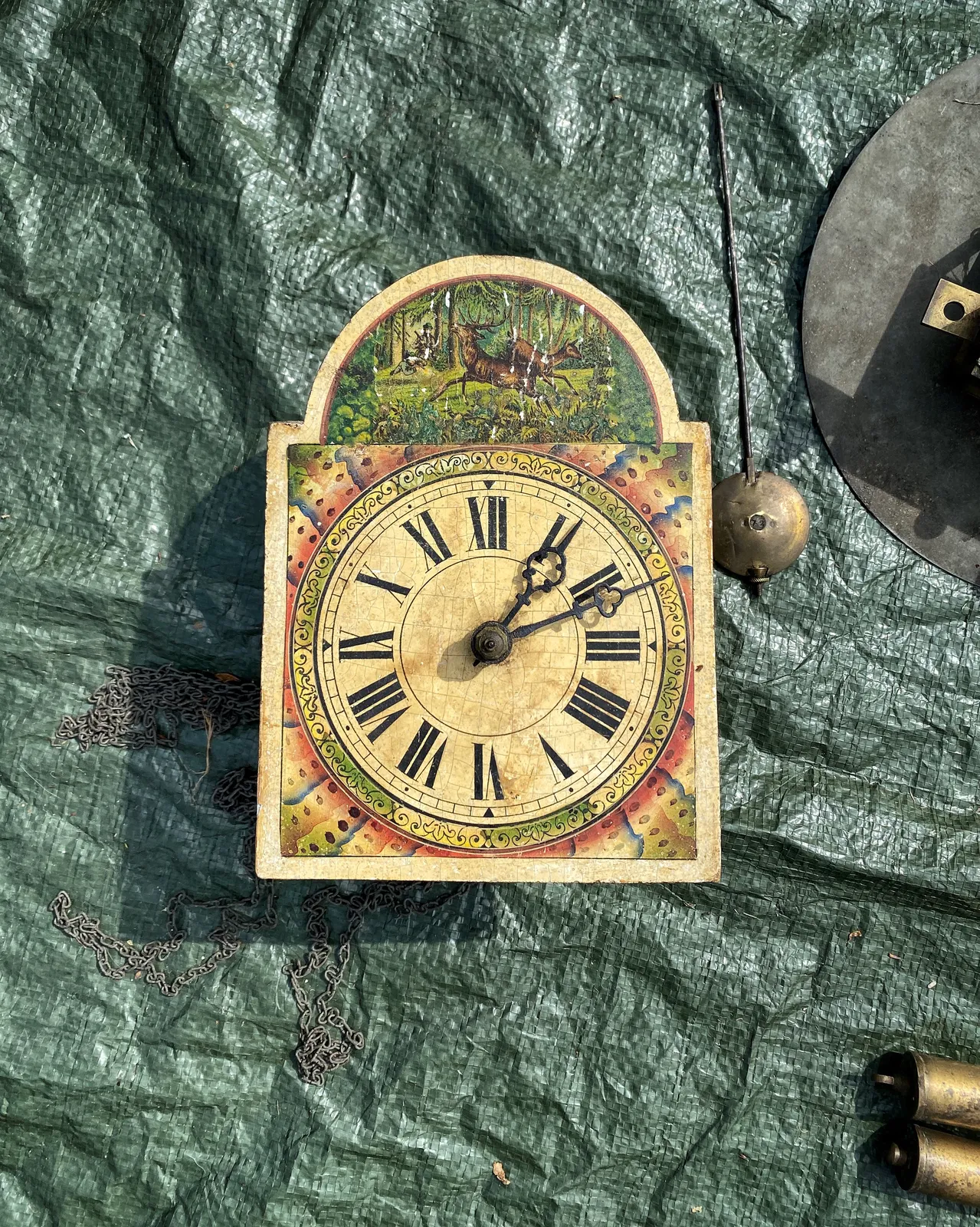A colourful, rather old clock lies on the ground. It’s deconstructed: the pendulum and the weights lay around it. The clock points at 11 past one.