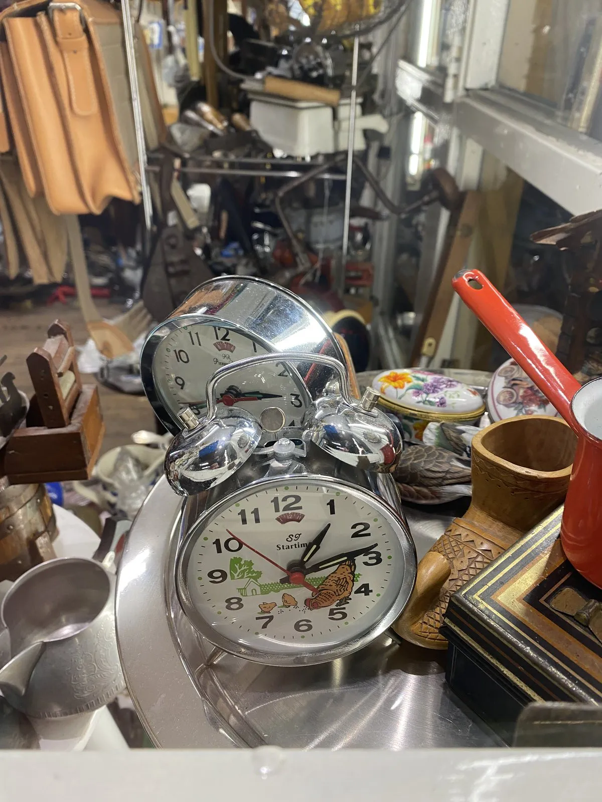 A picture of stuff in a thrift shop. There are two clocks, and the one in the foreground points at 13 past 1.