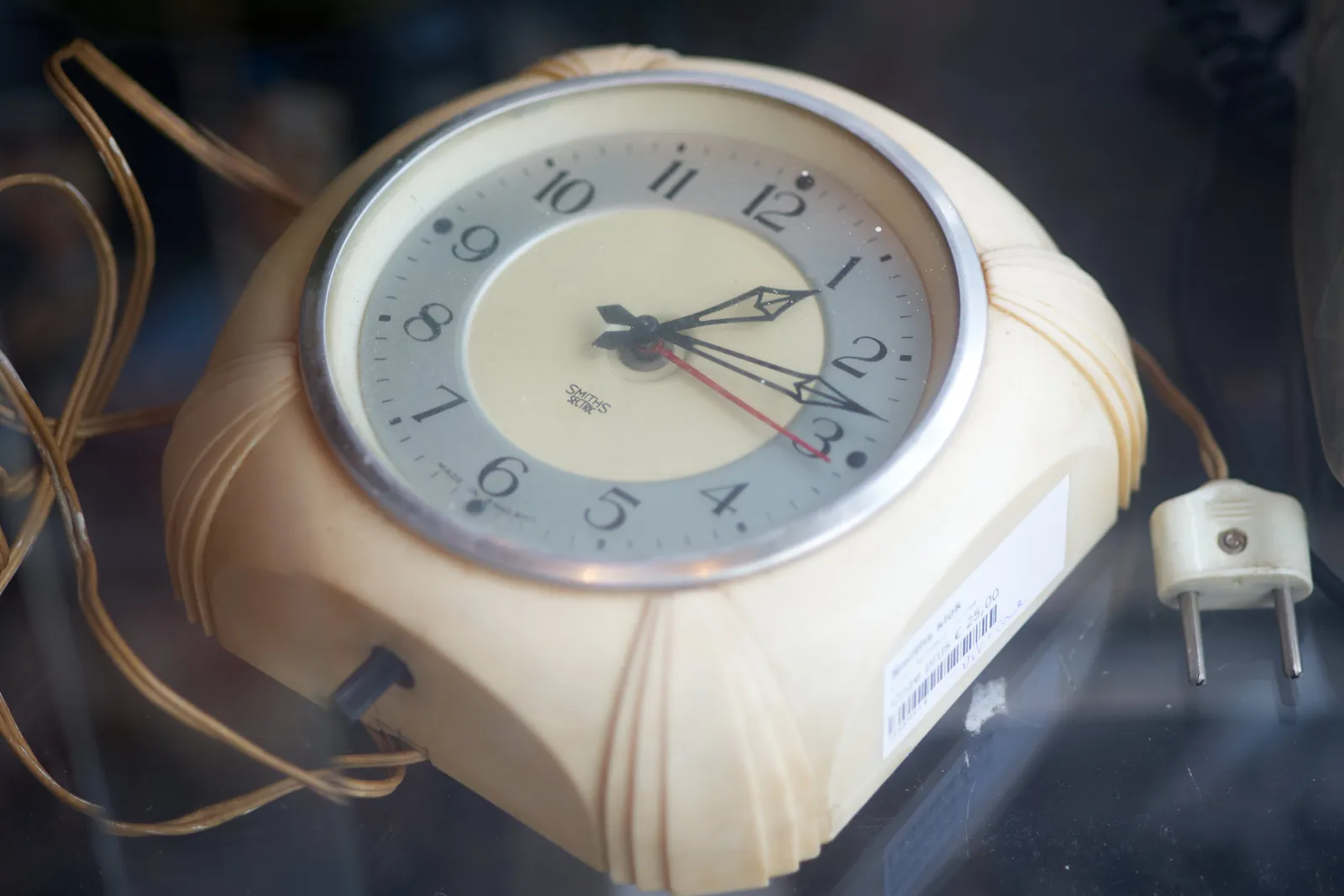 A plastic electric clock, with a long wire attached to it, unplugged, points at 13 past one. 