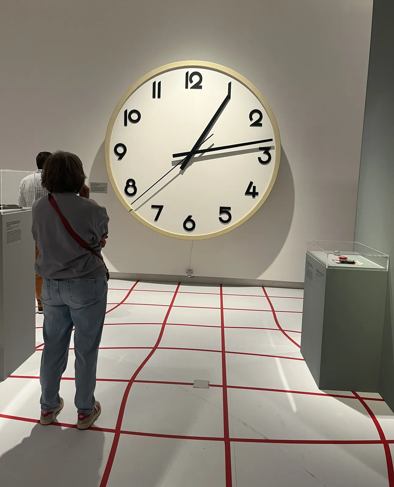 The red lines on this white floor are deliberately painted in non-straight lines, in order to form a wobbly grid. Two people are standing on this grid, looking at a huge wall clock. It points at 13 minutes past one.