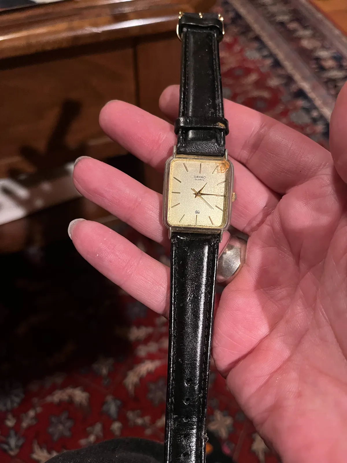 Someone is holding an old watch in their hand. In the background, blurry, an old Persian rug. The watch points at 14 minutes past one
