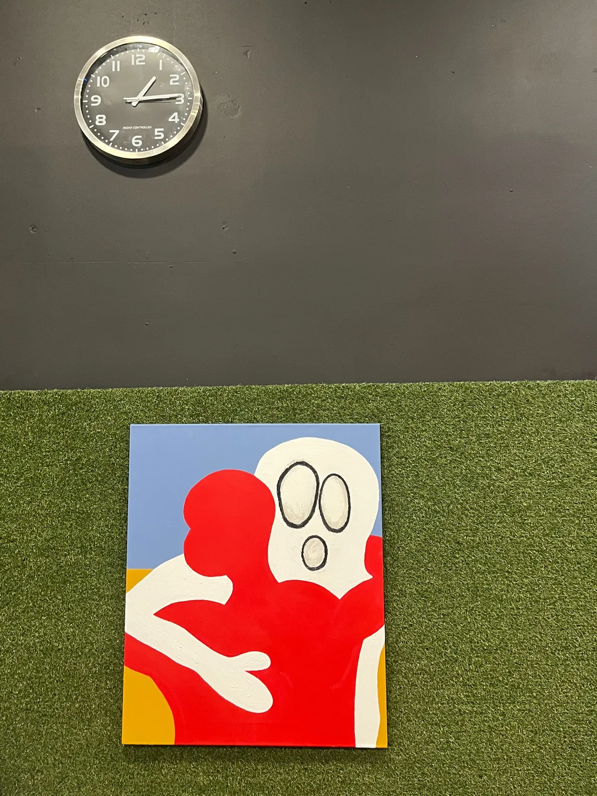 A painting of a red figure hugging a figure that reminds me of one the scream by Edvard Munch. The painting is hanging on a grasslike wall. Above this painting there’s a clock that points at 14 minutes past one.
