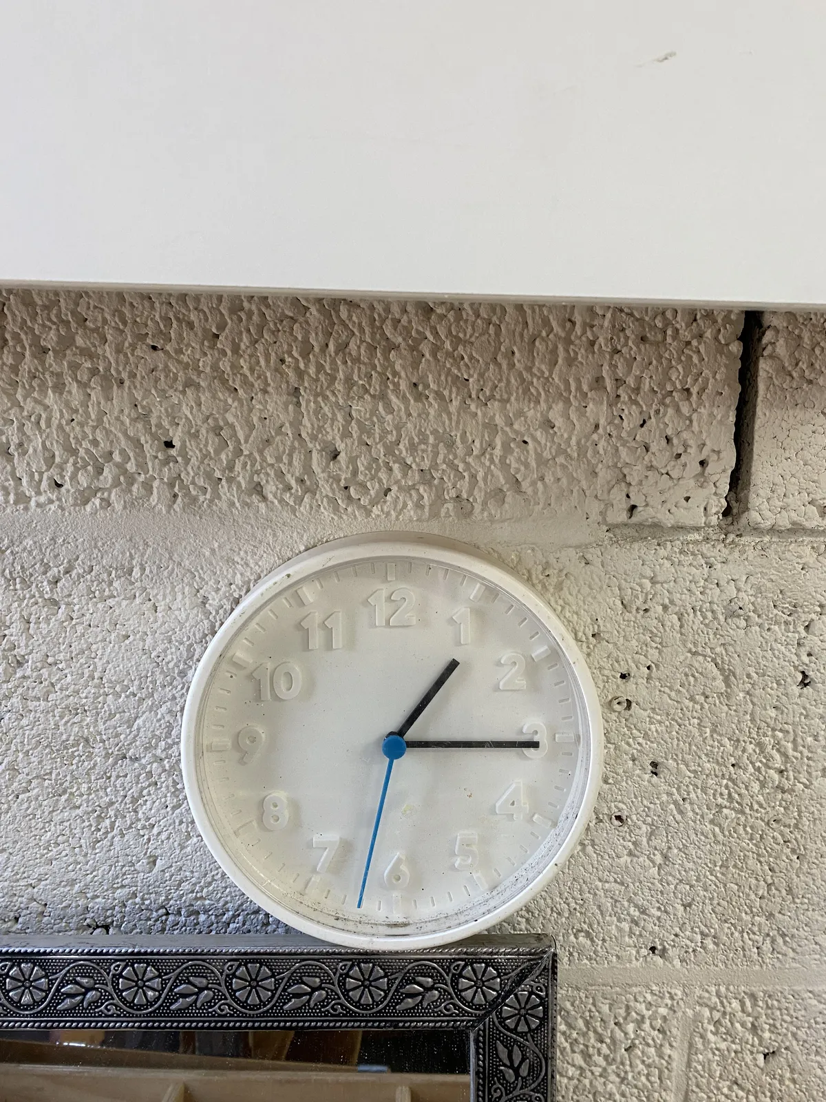 A white clock with the numbers and indicators embossed. The minute and hour hand are black, the second hand is blue. It points at a quarter past 1 and 32 seconds.