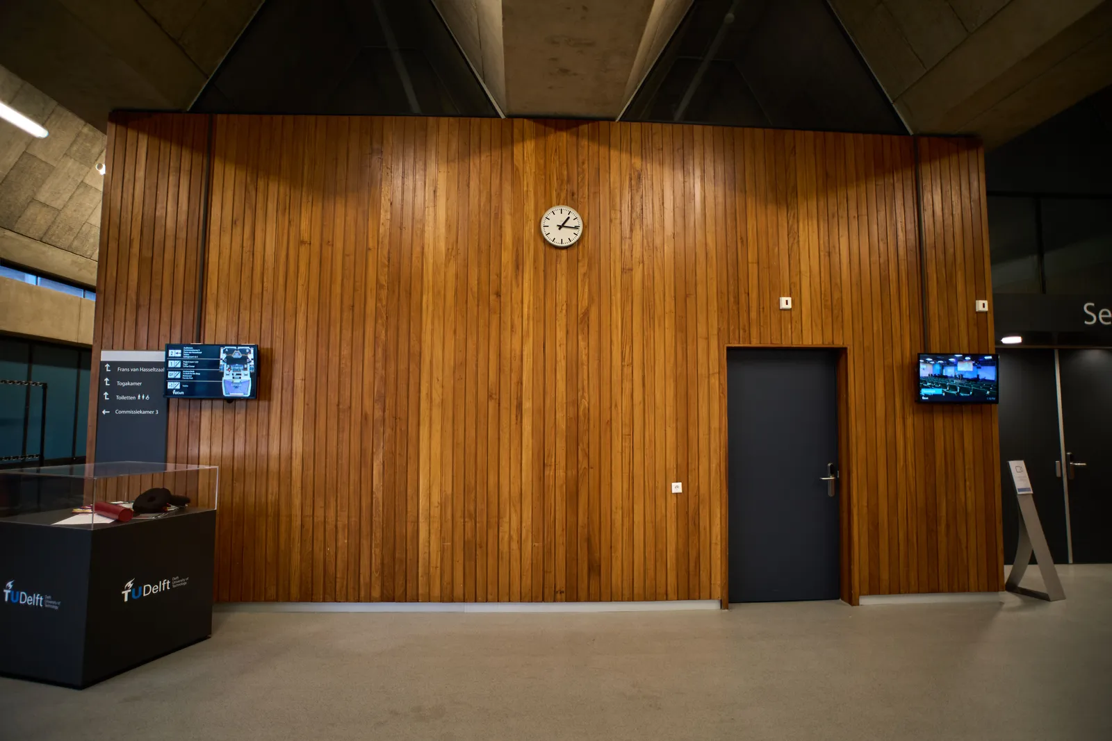A wall made from wooden laths. There’s a clock on it and it points at 16 minutes past one.
