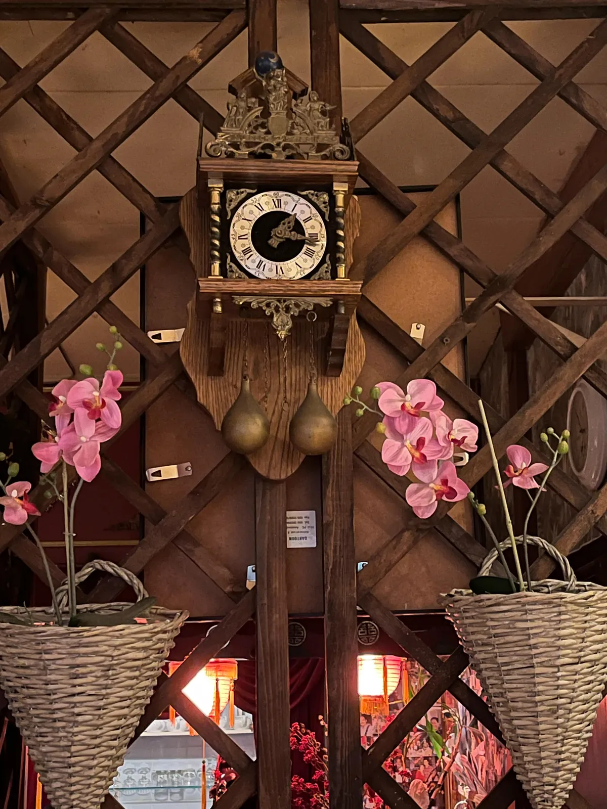 Quite a lot is going on in this picture. What seems to be a wall, turns out to be a wooden lattice with the back of a painting, and a part of a commercial kitchen behind it. In front of it are two wicker baskets, with flowers in them. Above these flowers there is an old cuckoo clock, which points at 16 minutes past one.  