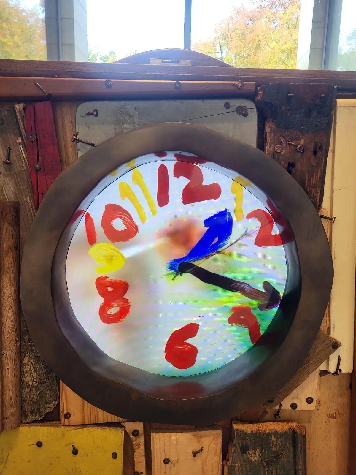 The numbers and the hands of this clock are painted onto glass with colourful paint. As if a child painted it. The clock seems to point at approximately 17 minutes past one. 